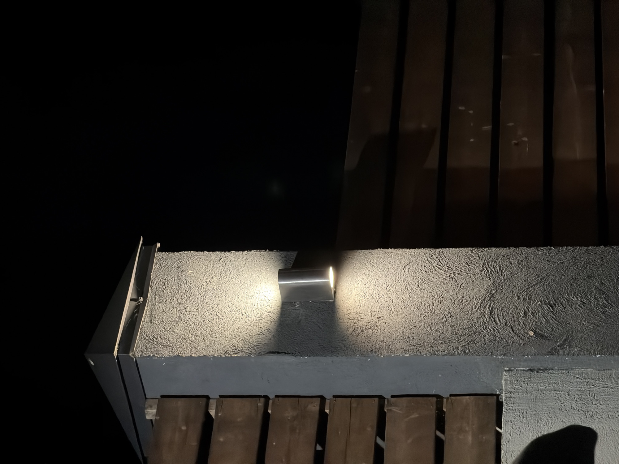 The image shows an outdoor pillar with a light fixture at night. The pillar is made of rough, textured concrete and has a flat, dark cap on top. A modern, cylindrical silver light fixture is mounted to the pillar, emitting light upwards and downwards, casting distinct shadows on the textured surface. To the left of the pillar, a section of a dark wooden fence is visible, constructed of horizontal planks. To the right, another dark wooden fence, also with horizontal planks, extends into the background. The overall scene is dark, with the light from the fixture being the primary source of illumination. The location is specified as Семилуки, Russia.