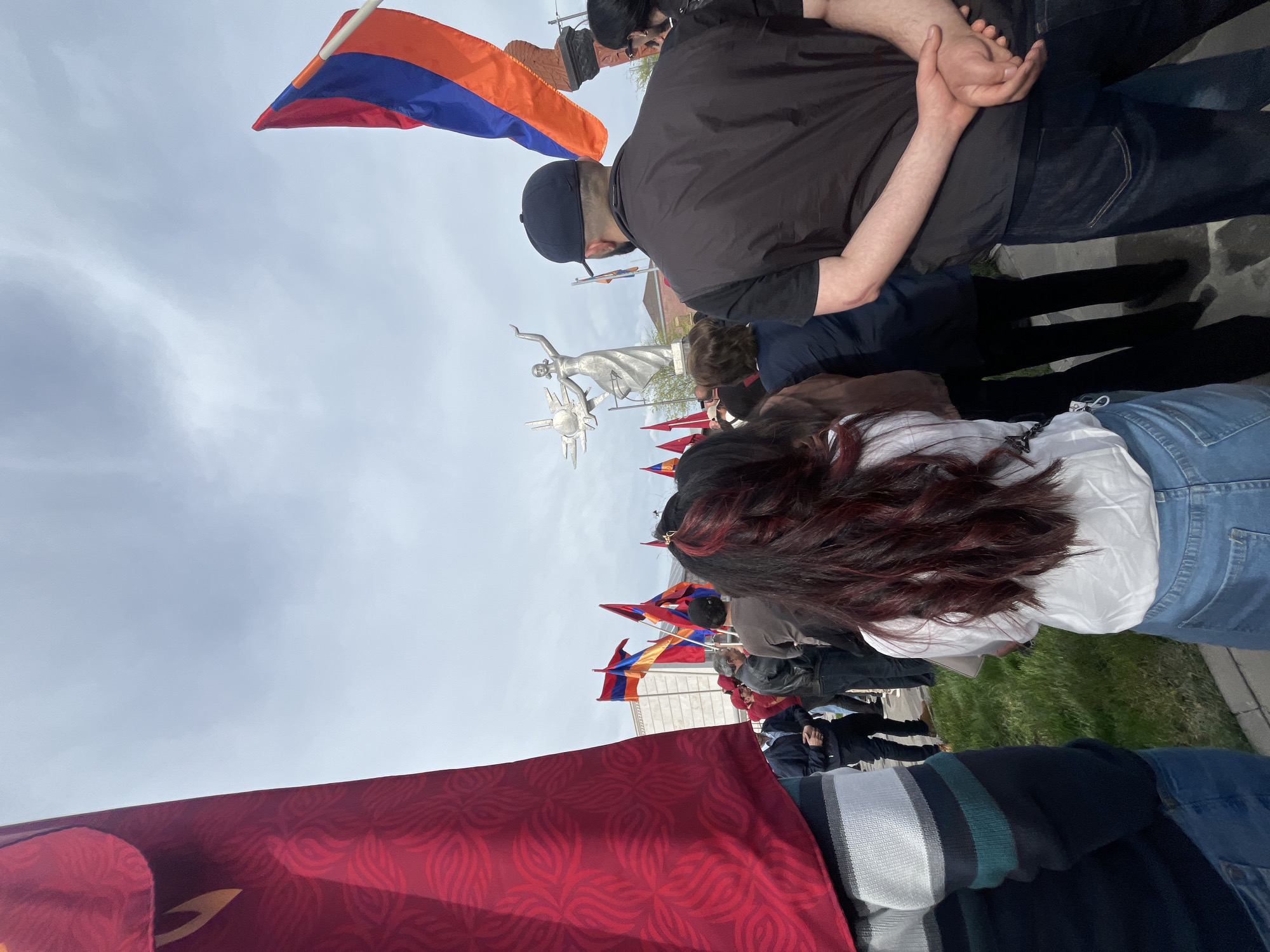 A low-angle, outdoor shot shows a gathering in what appears to be the city of Akhuryan, Armenia, based on the presence of the "Mother Armenia" monument visible in the middle distance. Several people are standing, holding or positioned near flags with a red, blue, and orange horizontal tricolor design.
In the foreground, the lower portion of a person wearing a striped sweater and denim jeans is visible. To their right, a large red flag with a patterned design is draped. In the middle of the frame, a woman with long dark hair that fades to red at the ends stands wearing a white shirt and blue jeans. Near her is a man with dark hair wearing a black cap and a black long-sleeve shirt with dark jeans. He has his hands clasped behind his back.
The silver "Mother Armenia" monument is visible against a pale sky. More flags are being held behind the monument. The lighting appears to be diffuse, suggesting an overcast day. The ground appears to be either paved or grassy.