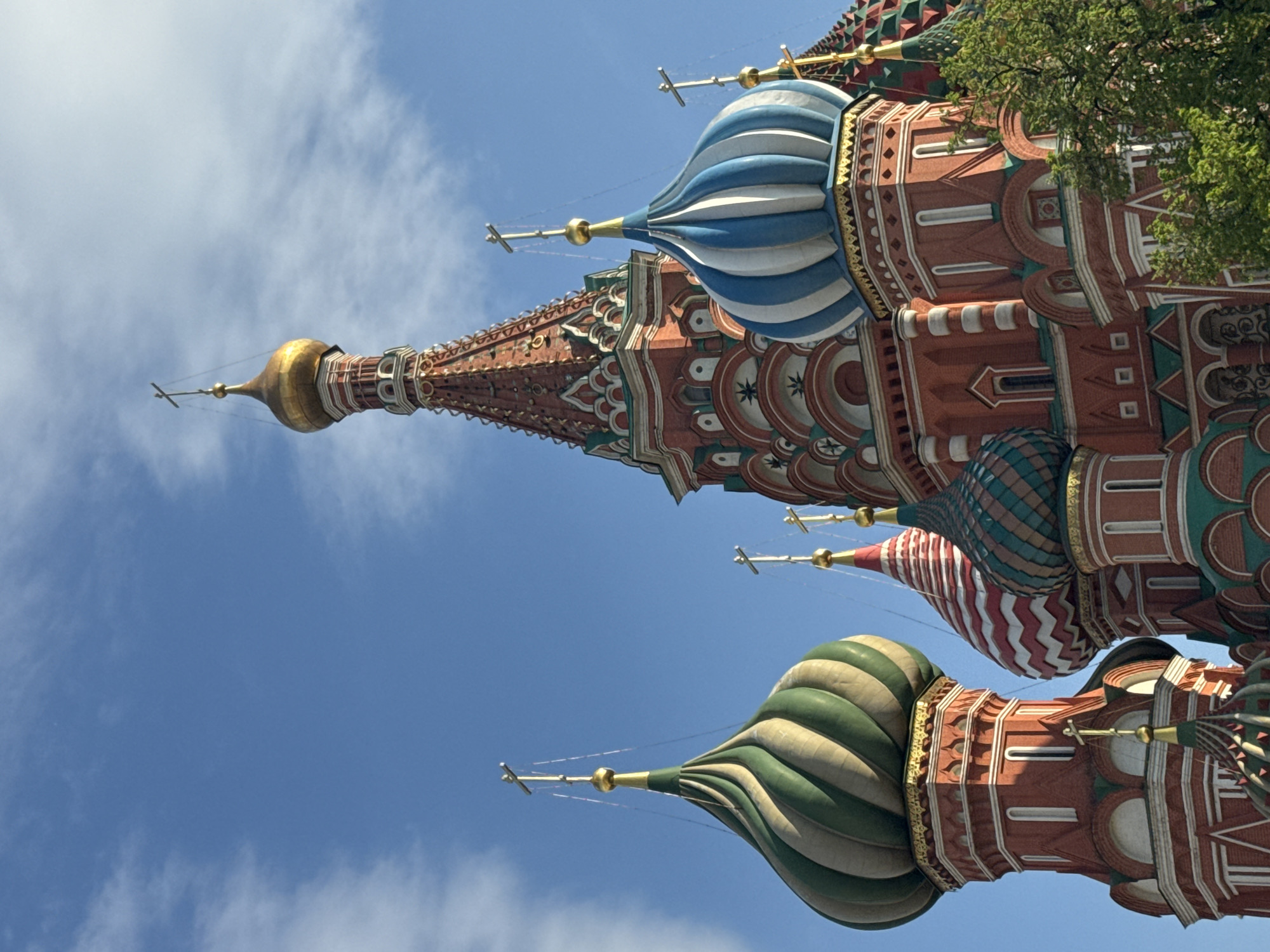 The image shows a portion of Saint Basil's Cathedral in Moscow, Russia, viewed from a low angle looking up. The cathedral's distinct onion-shaped domes are visible, each uniquely decorated with varying colors and patterns. The background consists of a bright blue sky with scattered white clouds. The architectural details of the cathedral are intricate, featuring red brickwork, decorative arches, and golden accents. Green foliage is present in the upper right corner of the frame, possibly from a nearby tree.