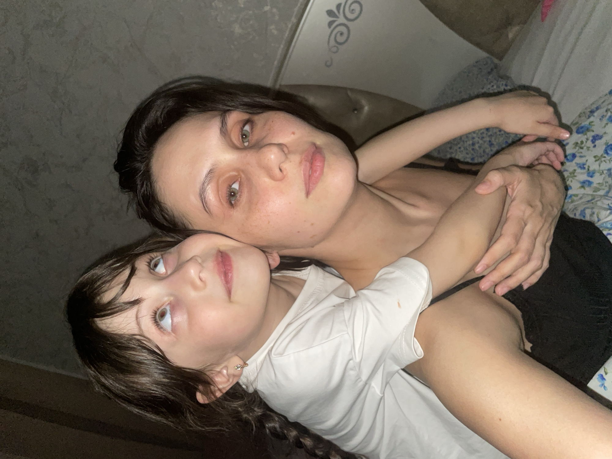 This image captures an intimate moment between an adult woman and a young girl, likely mother and daughter, embracing indoors. The photograph is oriented diagonally, taken from a slightly elevated perspective.
The adult woman is positioned behind the child, her head resting near the child's shoulder, and her arms wrapped around the girl. She has dark hair, light green/hazel eyes, and light skin with visible freckles, wearing a dark top. Her expression is calm, looking towards the viewer.
The young girl is in the foreground, partly facing away from the viewer, with her head tilted upwards. She has dark brown hair, light eyes, and light skin, wearing a white t-shirt and a small earring in her left ear. Her left arm is wrapped around the adult. Her gaze is directed upwards and to the side, suggesting contemplation or observation.
The background reveals a dark, textured wall or headboard with a subtle decorative swirl pattern on the left. On the right, blue floral patterned bedding is visible, suggesting a bed or a sofa. The lighting is dim, indicative of an evening or night-time setting. The overall mood conveys closeness, comfort, and affection between the two individuals. There is no visible text in the image. The location context is Izhevsk, Russia.