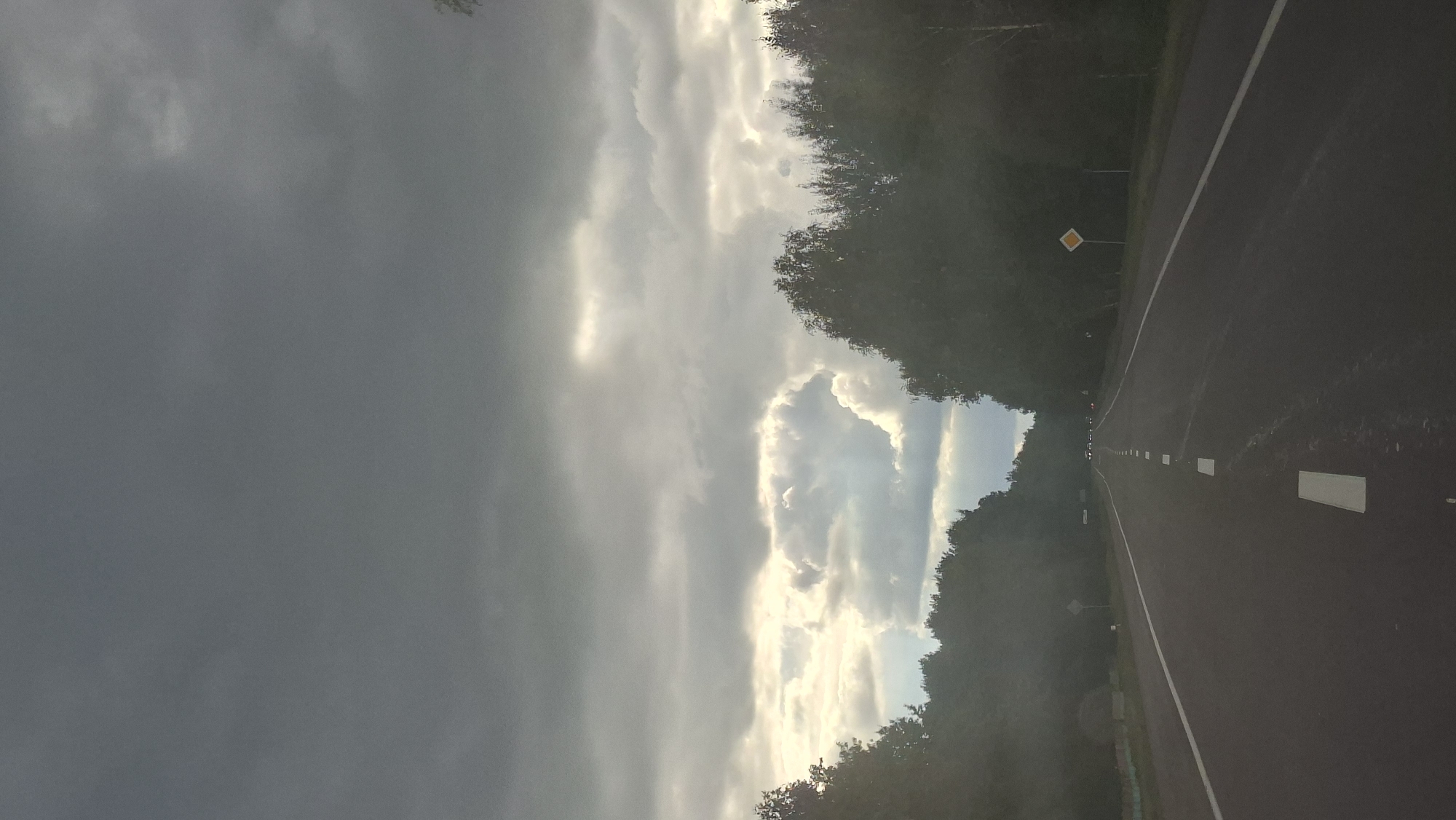 The image depicts a road with trees lining both sides. The sky is overcast with heavy, dark clouds, but there are breaks where sunlight is peeking through, creating bright patches. The road is asphalt and has white lane markings. There are no visible people or vehicles in the immediate foreground. In the distance, headlights from what appear to be several cars are visible on the road, suggesting traffic ahead. A diamond-shaped yellow road sign is present on the right side of the road. The overall scene suggests a somewhat gloomy or overcast day on a rural or semi-rural highway. The context provided places this scene in Астровенскі сельскі Савет, Belarus, implying a landscape typical of that region.