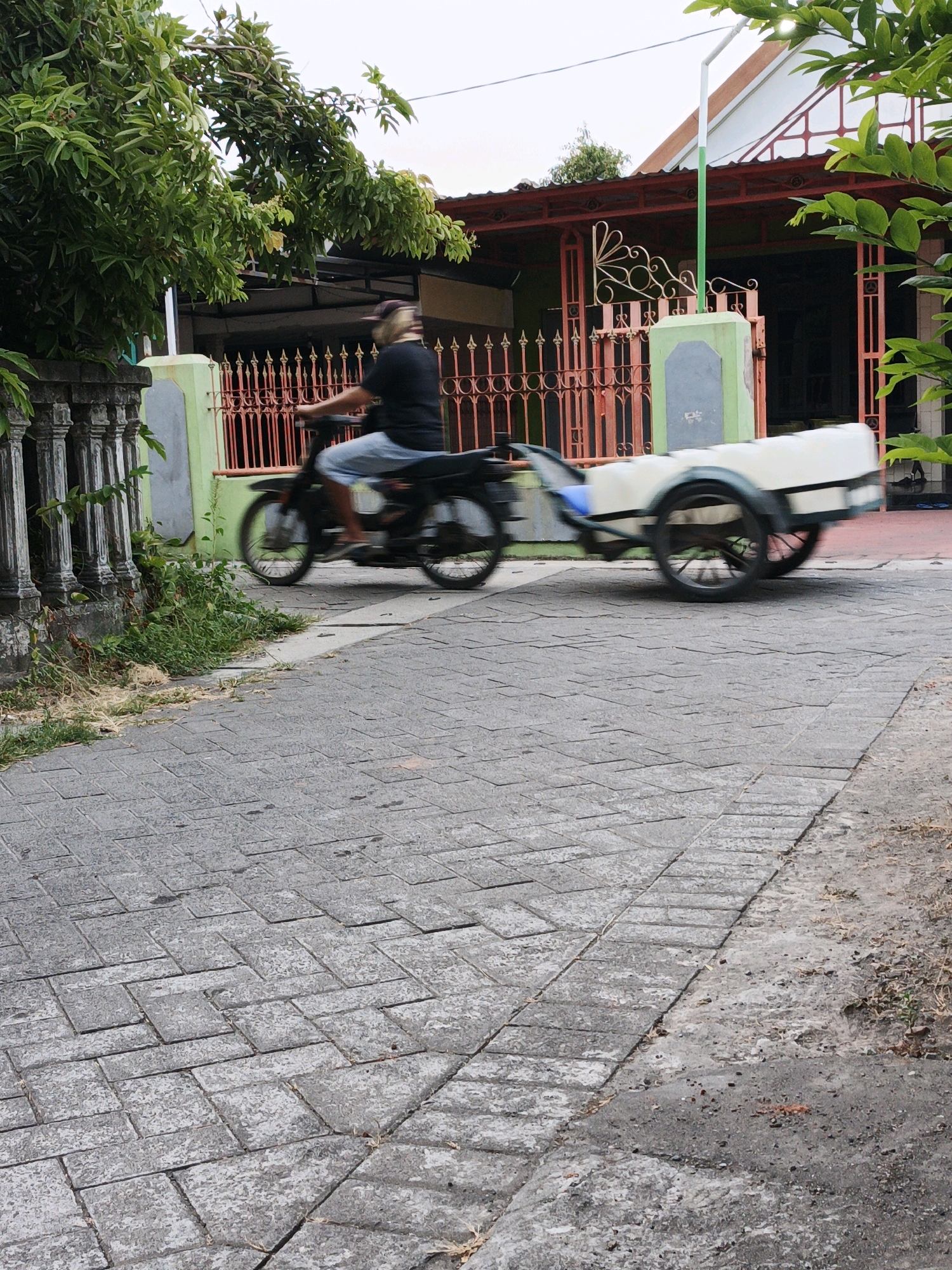 In Ketegan, Indonesia, a person rides a motorcycle with a trailer attached. The person, wearing a dark shirt and light shorts, is captured in motion, with the wheels of the motorcycle and trailer blurred. The motorcycle is a dark color, and the trailer is white with a green frame. They are traveling on a paved road made of interlocking gray bricks. To the left of the road, there is a weathered stone fence and overgrown greenery. Behind the fence, a building with a red roof and intricate orange railings is visible. The area is characterized by a mix of residential architecture and natural vegetation. The scene suggests a typical day in a suburban or semi-rural Indonesian setting.