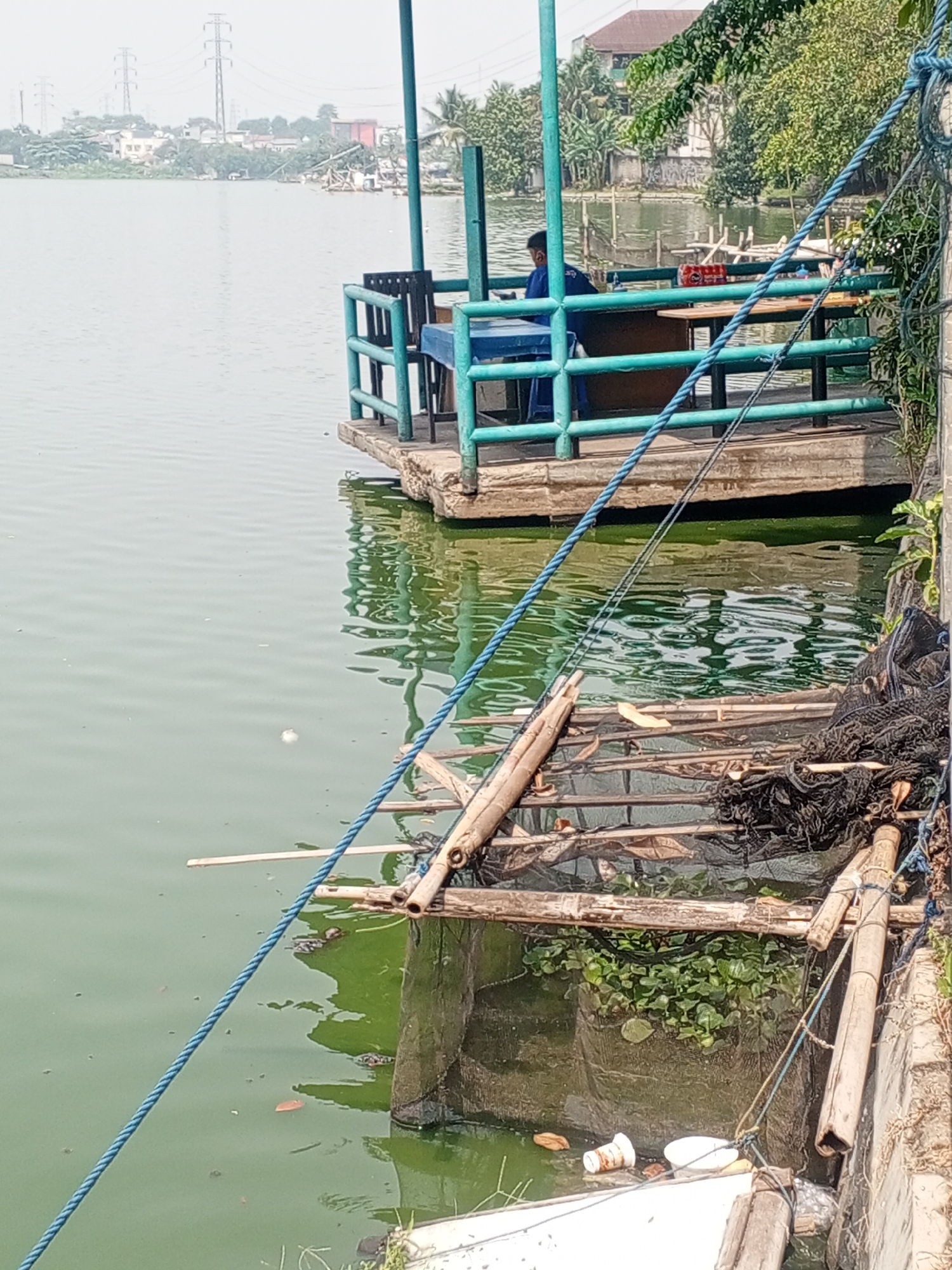 A person wearing a blue shirt sits at a table on a floating wooden deck. The deck has turquoise railings and is situated on a body of murky green water. In the background, there are trees, buildings, and electrical towers, indicating an urban or semi-urban environment. The foreground shows a close-up of the water's edge with bamboo structures, fishing nets, and some debris floating in the water. The scene appears to be in Depok, Indonesia, given the architectural styles and vegetation.