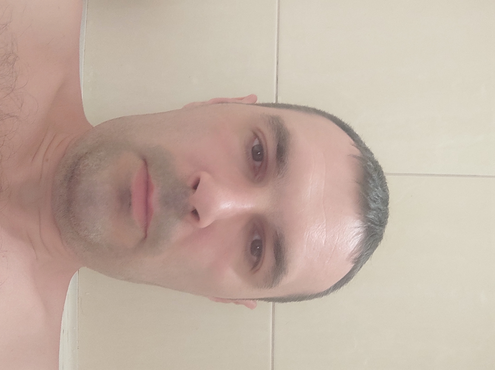 The image is a close-up, head-and-shoulders photograph of a light-skinned man with short, dark hair, a receding hairline, and a light stubble on his face. He has dark brown eyes and a neutral expression. The man is shirtless, revealing his shoulders and chest with some dark hair. The background consists of a light-colored, tiled wall. The lighting appears to be artificial and is slightly warm. There are no discernible landmarks or features that would indicate a specific location within the city of Kobeliaky, Ukraine. The photograph is taken from a frontal perspective, with the man's face filling most of the frame.