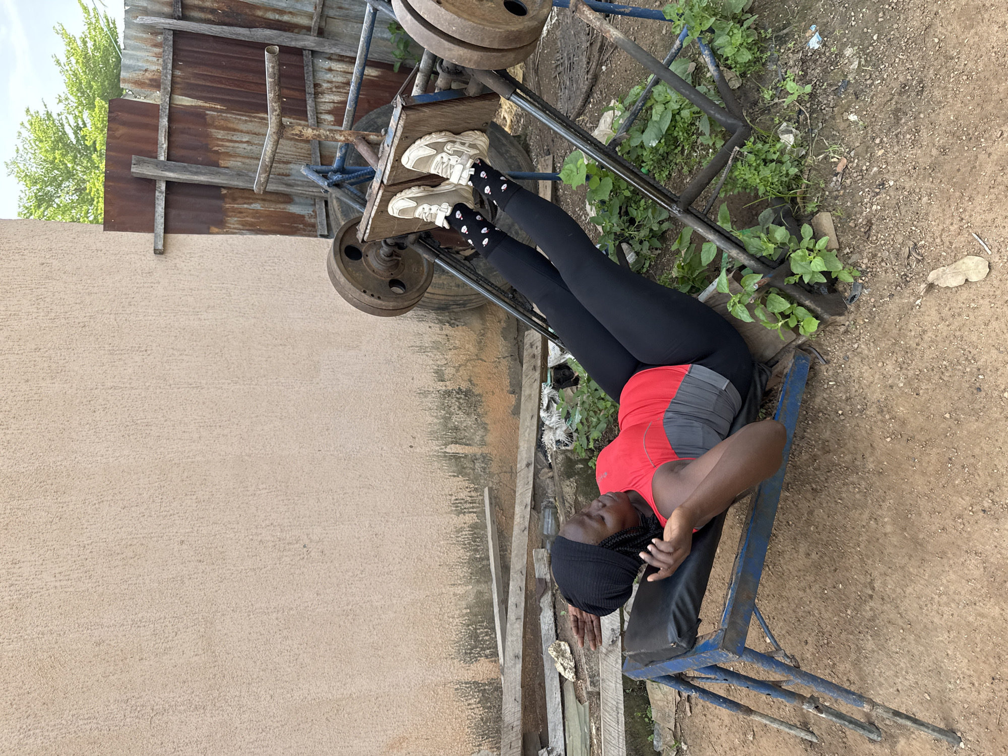 A young Black woman wearing a black headwrap, a red and gray tank top, black leggings, and white sneakers with black socks is lying on her back on a blue metal exercise bench. Her legs are extended upwards, with her feet resting on a wooden platform attached to a leg press machine. The machine is made of metal, with large circular weights attached to it, and appears to be outdoors. The background consists of a rough, light-colored wall, a rusty corrugated metal fence, and green foliage. The ground is dirt and gravel. The scene is likely located in a residential area or an outdoor gym in Akwanga, Nigeria.