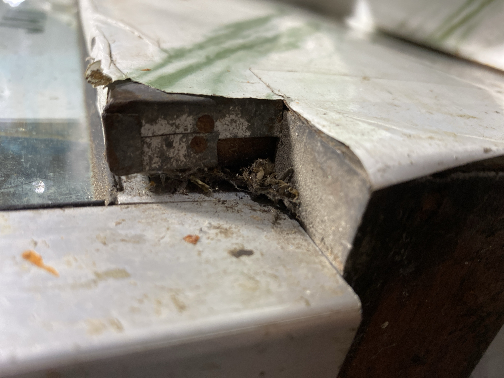 The image shows a close-up view of a dirty, weathered corner of what appears to be an outdoor window or door frame. There is dust, cobwebs, and debris accumulated in the crevice. The materials are a mix of metal and possibly wood, with peeling paint or coatings on the white surfaces. The lighting suggests it's daytime. The scene is devoid of people and any discernible activity. It is an urban scene, possibly in a residential or commercial area, with the location being Kasihan, Indonesia.