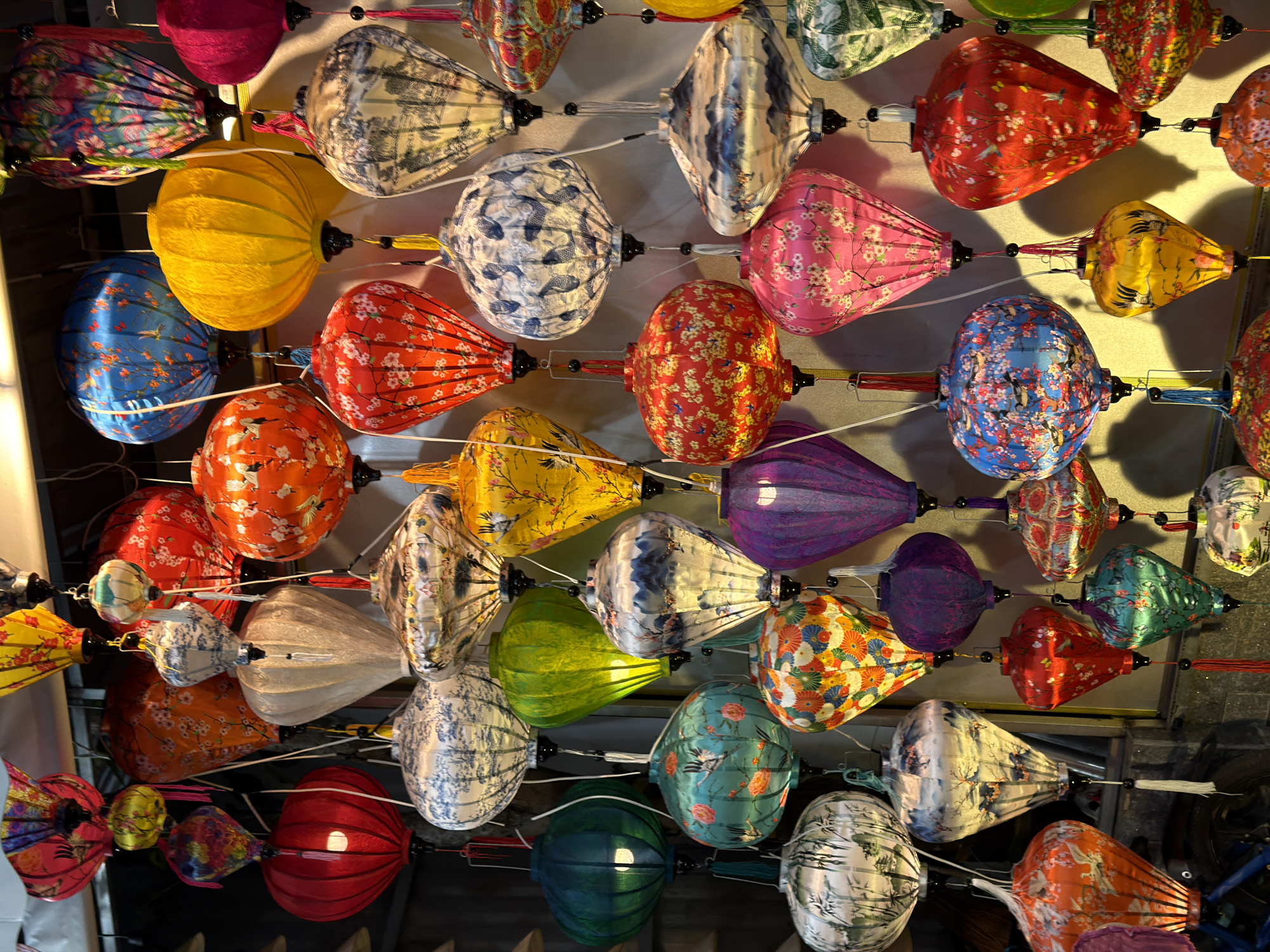 The image depicts a variety of colorful, decorative lanterns hanging in a shop, likely in Hội An, which is near Đà Nẵng, Vietnam. The lanterns vary in shape, size, and design, showcasing different patterns, colors, and fabrics. They are suspended from a ceiling or display structure. No people are visible in the frame. The environment is contained within a retail setting.