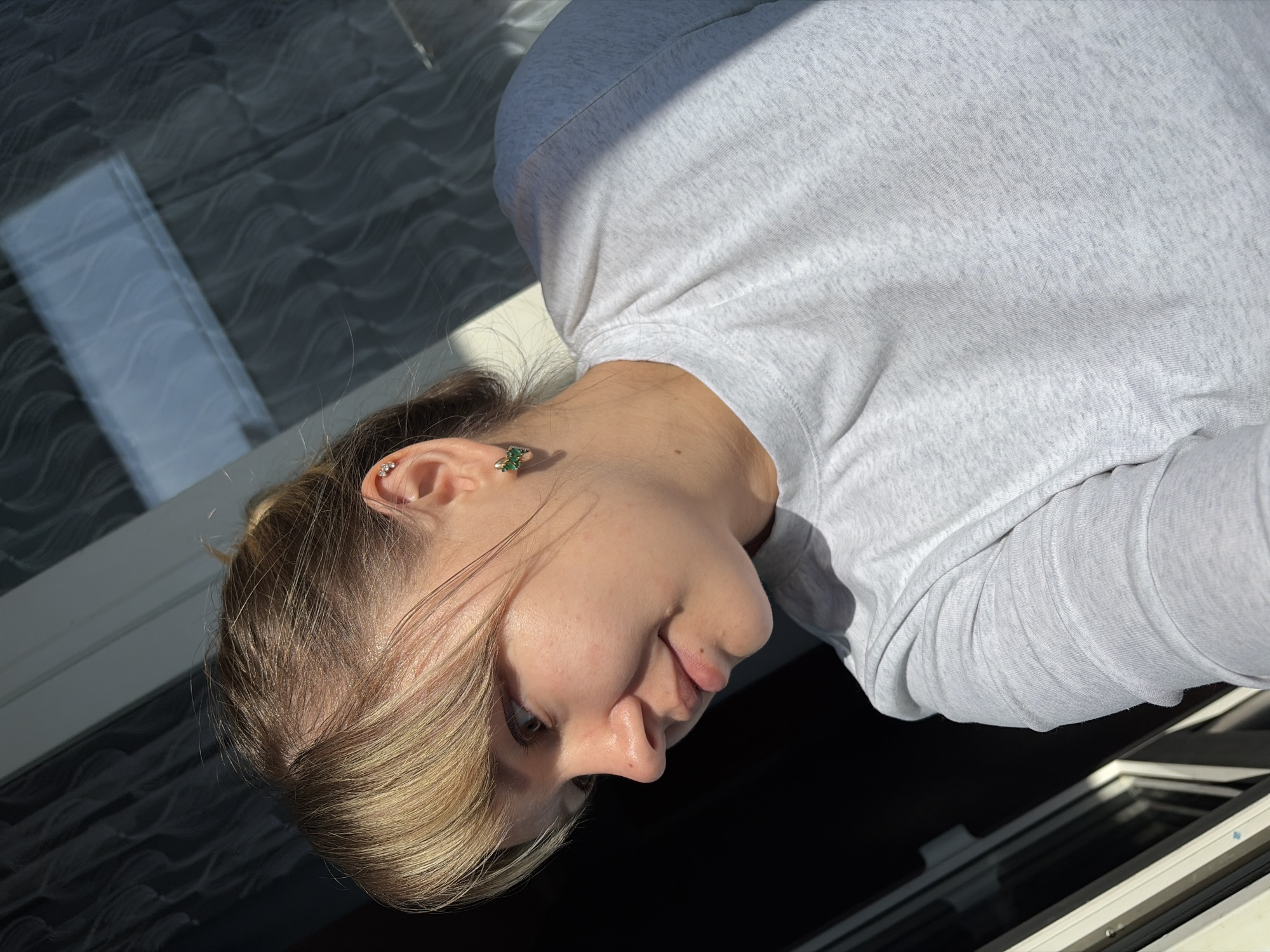 A young woman with blonde hair, wearing a light grey long-sleeved shirt, is looking down at an open window. She has a green earring in her left ear and another stud piercing higher up. The scene appears to be indoors, judging by the reflections in the window and the interior window frame. There's no indication of Chaikovsky, Russia, in the image.