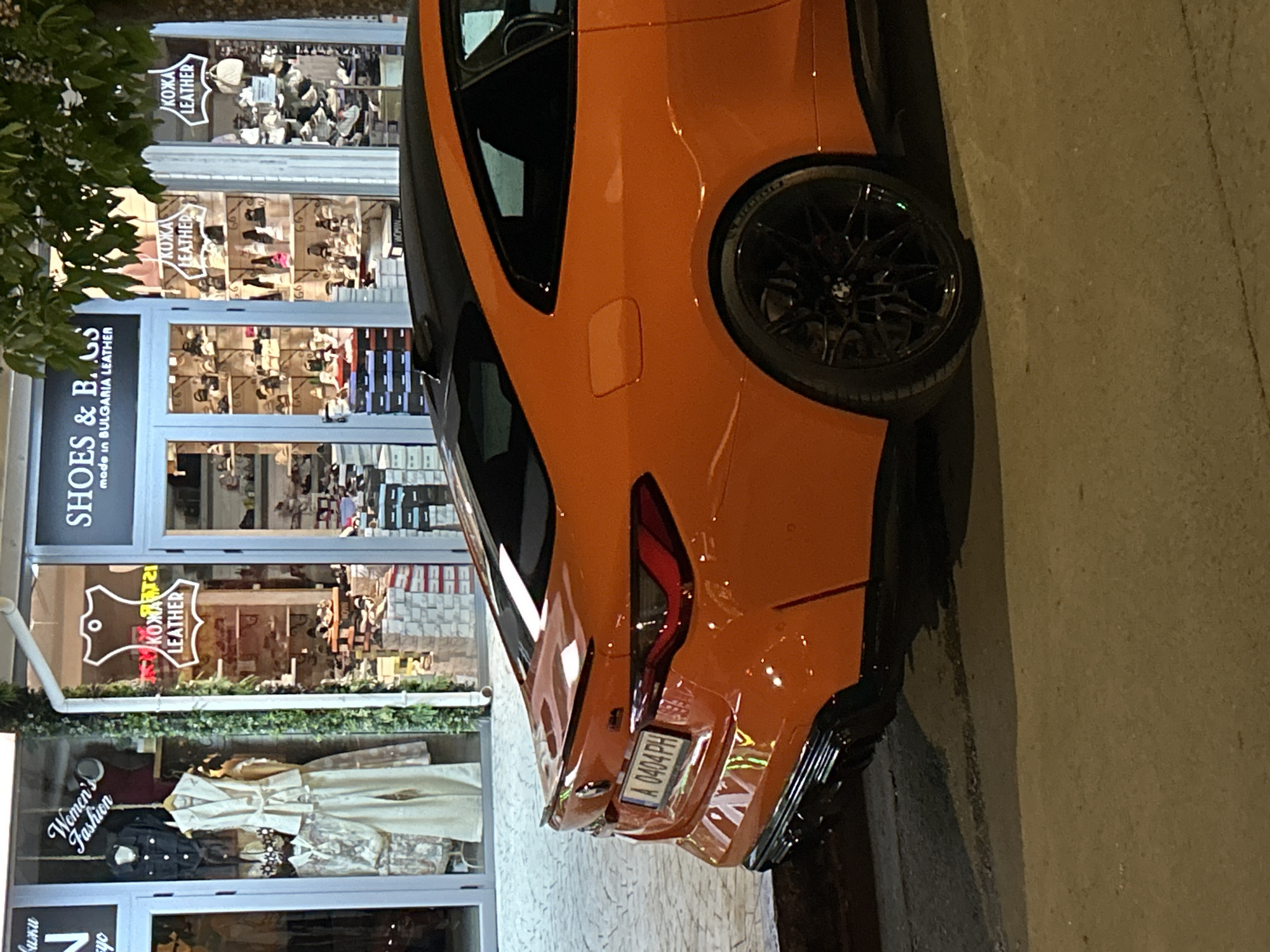 The image depicts the rear of an orange BMW M4 parked on a sidewalk in Nesebar, Bulgaria, facing a storefront. The storefront has several sections, displaying clothing, shoes, and leather goods. Signs read "Women's Fashion," "SHOES & BAGS made in BULGARIA LEATHER," and "KOЖA LEATHER" in both Cyrillic and English. The car's license plate is visible, reading "A 0404 PH." Vegetation is visible on the left of the frame. The ground surface is concrete. The photo was taken at night, as indicated by the lighting and reflections in the storefront windows.