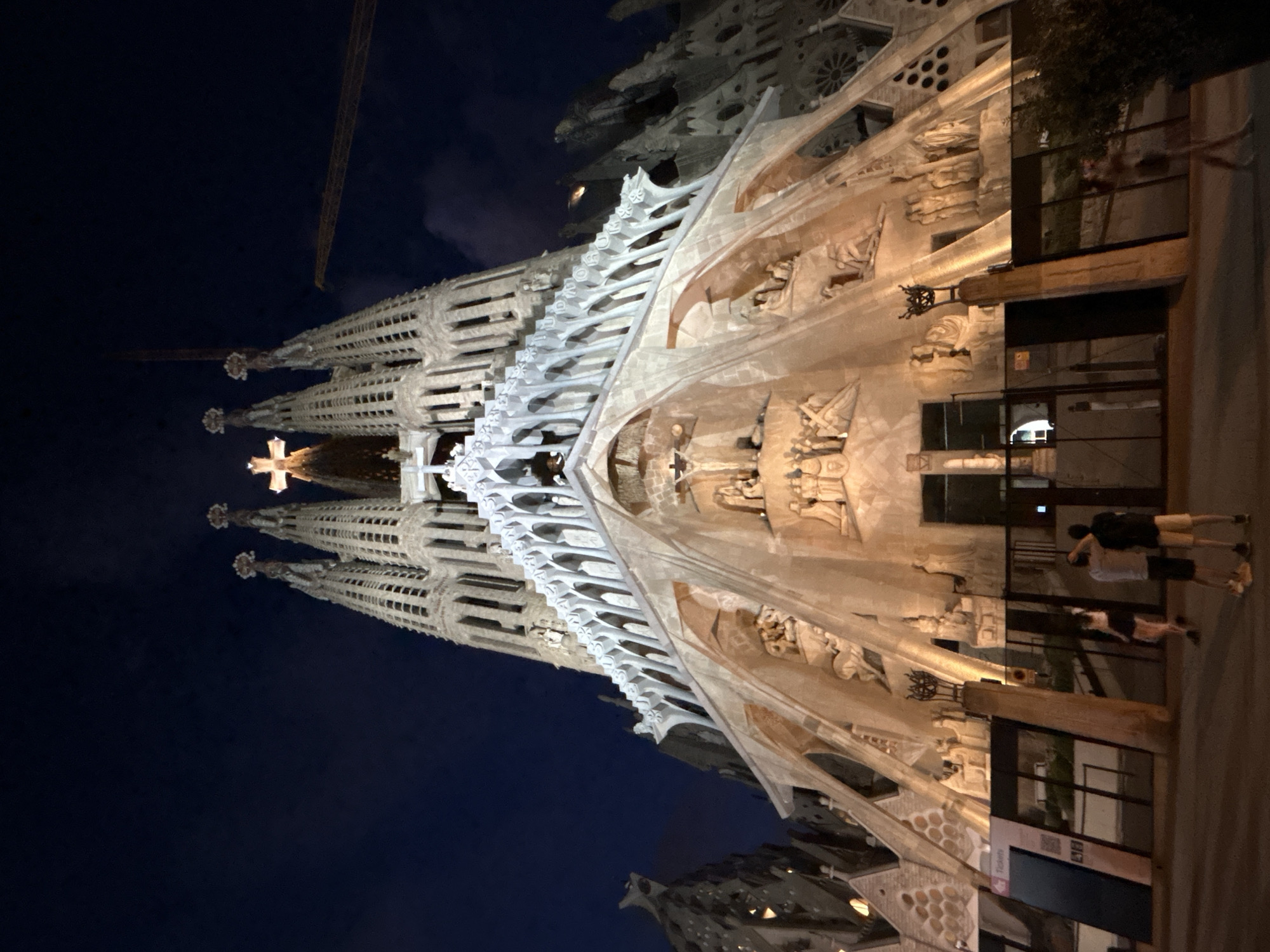 The image shows the Sagrada Familia basilica in Barcelona, Spain, at night. The iconic basilica, designed by Antoni Gaudí, is illuminated, highlighting its intricate facade and towering spires. The sky above is a deep blue, suggesting it is nighttime. In the foreground, a few people are visible, appearing to be tourists, standing and observing the basilica. One person is taking a photo. There are also some fences and signage in the foreground. In the upper right corner of the image, a construction crane is visible, indicating ongoing construction or renovations. The overall scene is one of awe-inspiring architecture and a quiet evening in a bustling city.