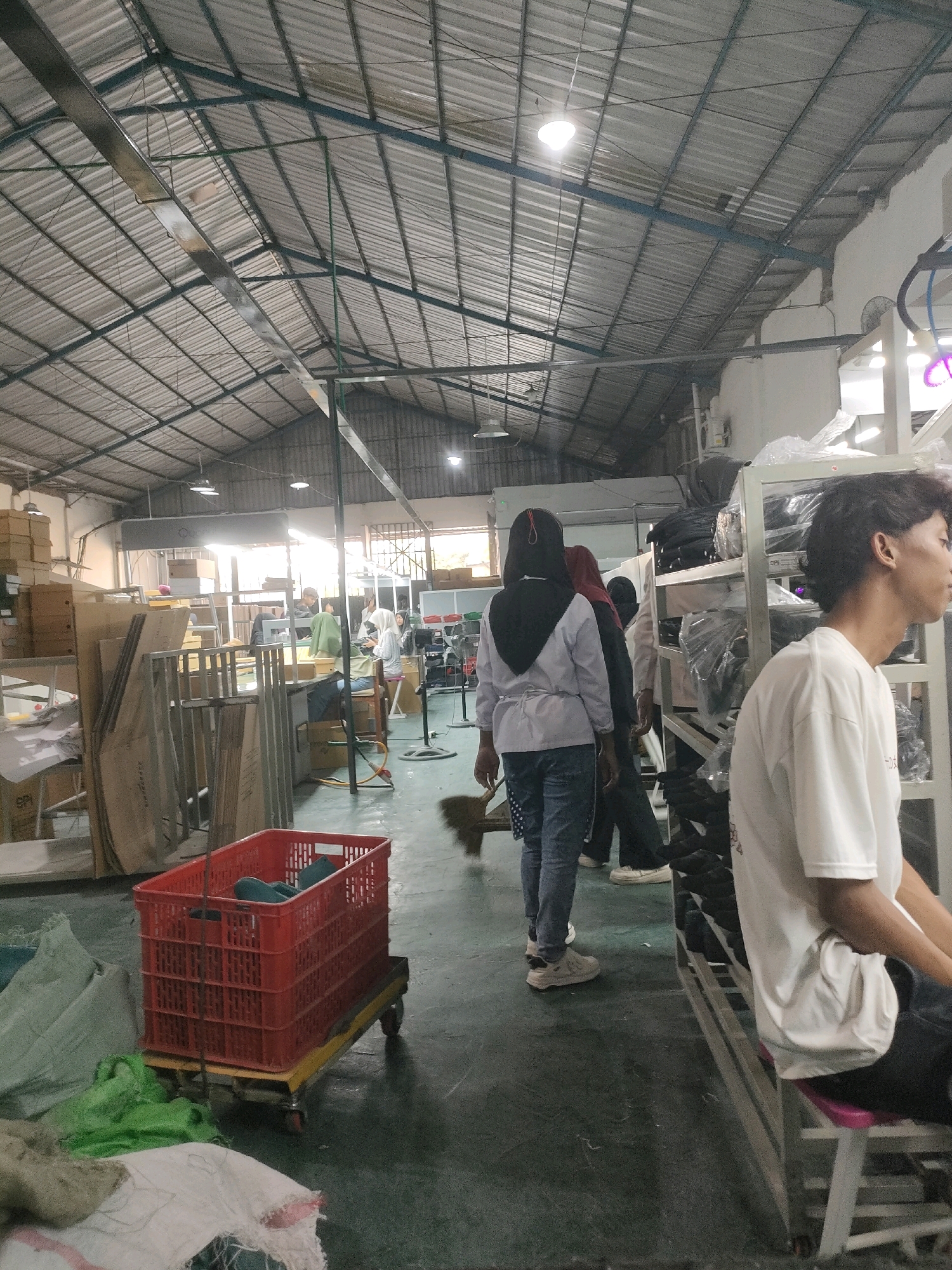 The image depicts the interior of a warehouse or factory setting in Trangsan, Indonesia. The ceiling is constructed of corrugated metal panels supported by a network of metal beams and trusses. Fluorescent lights are visible, providing illumination. The floor is a dark, worn concrete surface.
In the foreground, a red plastic crate rests on a yellow wheeled cart. Several large sacks, some green and white, are piled near the cart.
Several individuals are present. A young man in a white t-shirt is seated on a pink stool, facing away from the camera, and appears to be looking to his right. Further into the scene, a woman wearing a light-colored jacket and jeans stands with her back to the camera, holding a broom. She is accompanied by another person wearing a hijab and a third person partially visible behind her. Other individuals can be seen in the background, some appearing to be working at tables or stations.
Various objects suggestive of manufacturing or storage are present, including stacks of cardboard boxes, metal shelving units filled with items wrapped in plastic, and what appear to be raw materials or finished products. The overall environment suggests an active workspace.