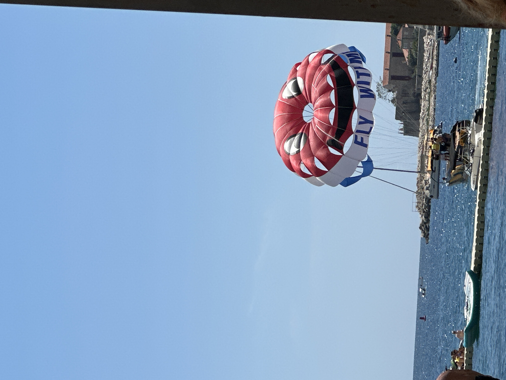 The image shows a parasail with a cartoonish face design, red, white, and blue in color, with the words "FLY WITH ME" printed on it. It is suspended in a clear blue sky over the Adriatic Sea. Below the parasail, a boat is visible, with several people on it, presumably waiting to go parasailing. The sea is a deep blue, with some waves. In the background, there is a rocky coastline with buildings, indicative of a coastal town. This scenery is characteristic of Budva, Montenegro, a popular tourist destination known for its beaches and recreational activities on the sea. The lighting suggests it is a sunny day.