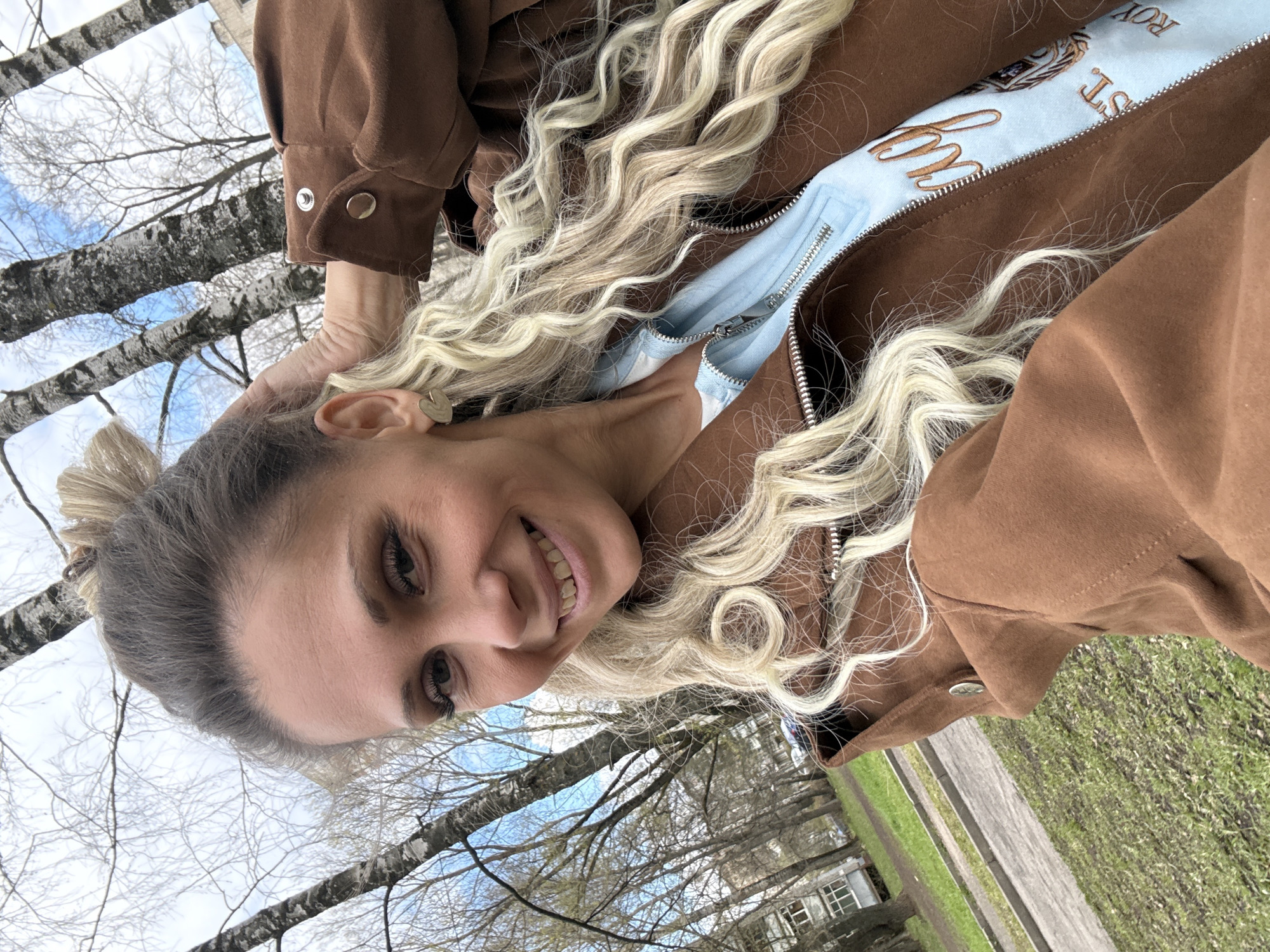 The image is a rotated selfie portrait of a young woman taken outdoors in Rusanovo, Russia. The woman is smiling broadly, looking directly at the camera, with her head slightly tilted. Her long, wavy blonde hair flows over her shoulders and chest, and she is holding some strands near her left ear, which has a small, gold-colored heart-shaped earring.
She is wearing a brown zippered jacket or hoodie with visible snap buttons on the cuffs. Underneath, a light blue zippered top is visible, featuring embroidered text. The visible text includes "ROYAL" (partially obscured), "ST.", and a cursive word that appears to be "My" or similar.
The outdoor setting includes several bare-branched trees with light-colored bark, possibly birches, against a bright, partly cloudy blue sky. In the midground and background, there are patches of green grass, a paved path or road, and a distant building with windows. The scene suggests a cool but bright day, possibly early spring or late autumn. The image is oriented such that the woman's head is on the left side of the frame.
