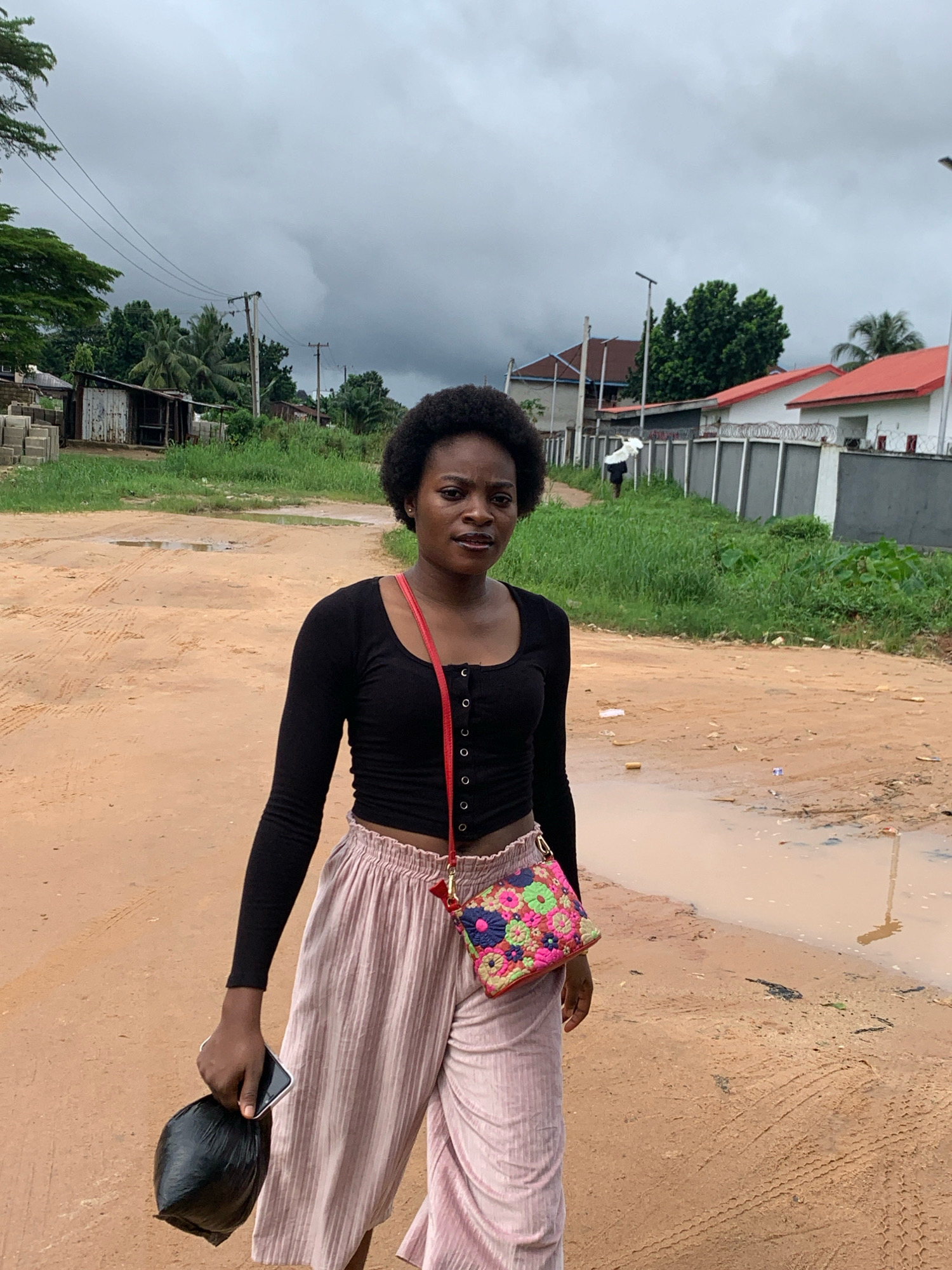 A young woman with dark, curly hair and medium-brown skin stands in the middle of a dirt road, looking directly at the camera. She is wearing a black, long-sleeved crop top with a lace-up detail on the front and loose, light pink, striped pants. A colorful, embroidered crossbody bag with floral patterns is slung over her right shoulder. In her left hand, she holds a black plastic bag and a white mobile phone. The road is unpaved and appears to be wet in places, with several puddles visible. The background shows a residential area with various buildings, including houses with red roofs and a smaller, dilapidated structure. Green vegetation and trees are scattered throughout the scene, and utility poles with wires are visible. The sky is overcast and cloudy. The scene is consistent with a rural or semi-urban environment in Umuode, Nigeria, likely during or after a rainfall.