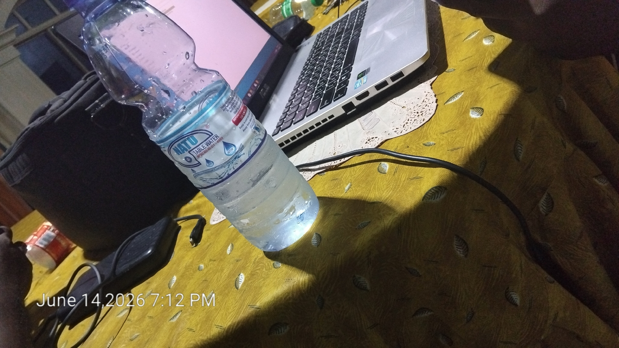 A partially filled bottle of NATO brand table water stands on a yellow tablecloth with a leaf pattern. A silver laptop with a black keyboard is open to the right of the water bottle. A black laptop bag is visible to the left of the bottle. A black cable extends from the laptop toward the bottom of the frame. The bottom left corner of the image shows a timestamp "June 14, 2026 7:12 PM". The scene appears to be indoors.