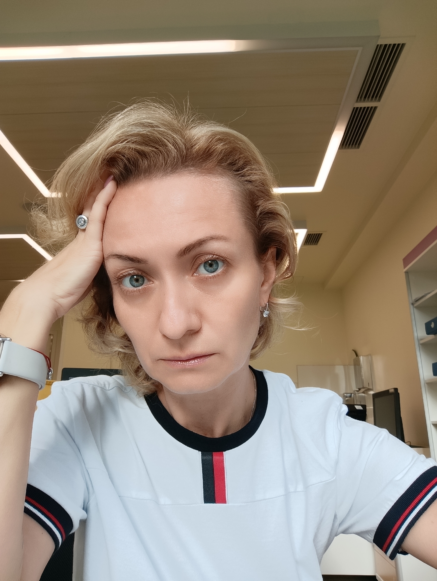 A close-up, frontal selfie of a blonde woman in her late 30s or early 40s with bright blue eyes. She has her right hand resting on her forehead and is wearing a white t-shirt with black, red, and white striped trim on the collar and sleeves. She has a silver ring on her right ring finger and is wearing a silver smartwatch on her left wrist. Her hair is short and styled in loose waves. The background appears to be an indoor office or modern waiting room with beige walls and ceiling panels with integrated lighting. There are also air vents visible on the ceiling. The lighting is bright and even. The image is taken from a slightly low angle. There is no identifiable information about the specific location in Moscow.