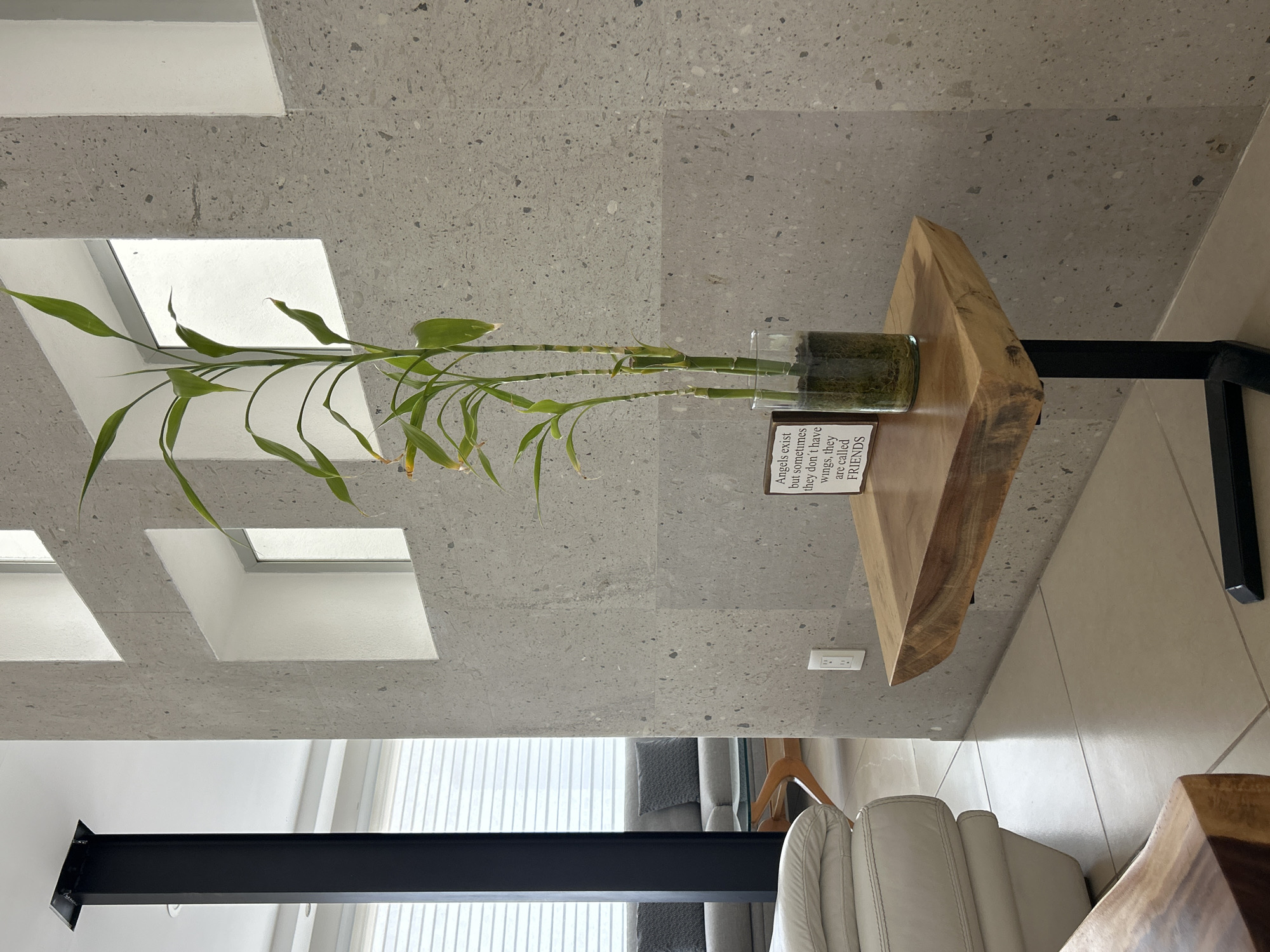 This is an indoor, well-lit photograph showcasing a modern interior design with a focus on a decorative plant. The main subject is a lucky bamboo plant, with multiple green stems and leaves, housed in a clear glass vase. The vase contains green material, possibly decorative stones or gel beads, at its base. The plant sits atop a small, natural-edge wooden side table with a black metal base.
To the left of the plant on the table, a small white rectangular plaque is visible, bearing the text: "Angels exist but sometimes they don't have wings, they are called FRIENDS".
The background wall is composed of light grey terrazzo or concrete tiles, flecked with darker and lighter aggregate. Several square, recessed openings are cut into this wall, adding an architectural detail. In the lower portion of the image, a large window with vertical white blinds is partially visible, suggesting natural light is illuminating the scene, indicating it's daytime. Below the table, a white electrical outlet is mounted on the wall. A section of a cream or light-colored upholstered armchair is also visible in the lower right, and a prominent black beam runs across the lower left, possibly a structural element or part of a furniture piece. The floor appears to be a light, neutral-toned tile.
The overall scene conveys a sense of calm, modern aesthetics, with the plant adding a touch of nature and the plaque providing a heartfelt message. There is no activity taking place.