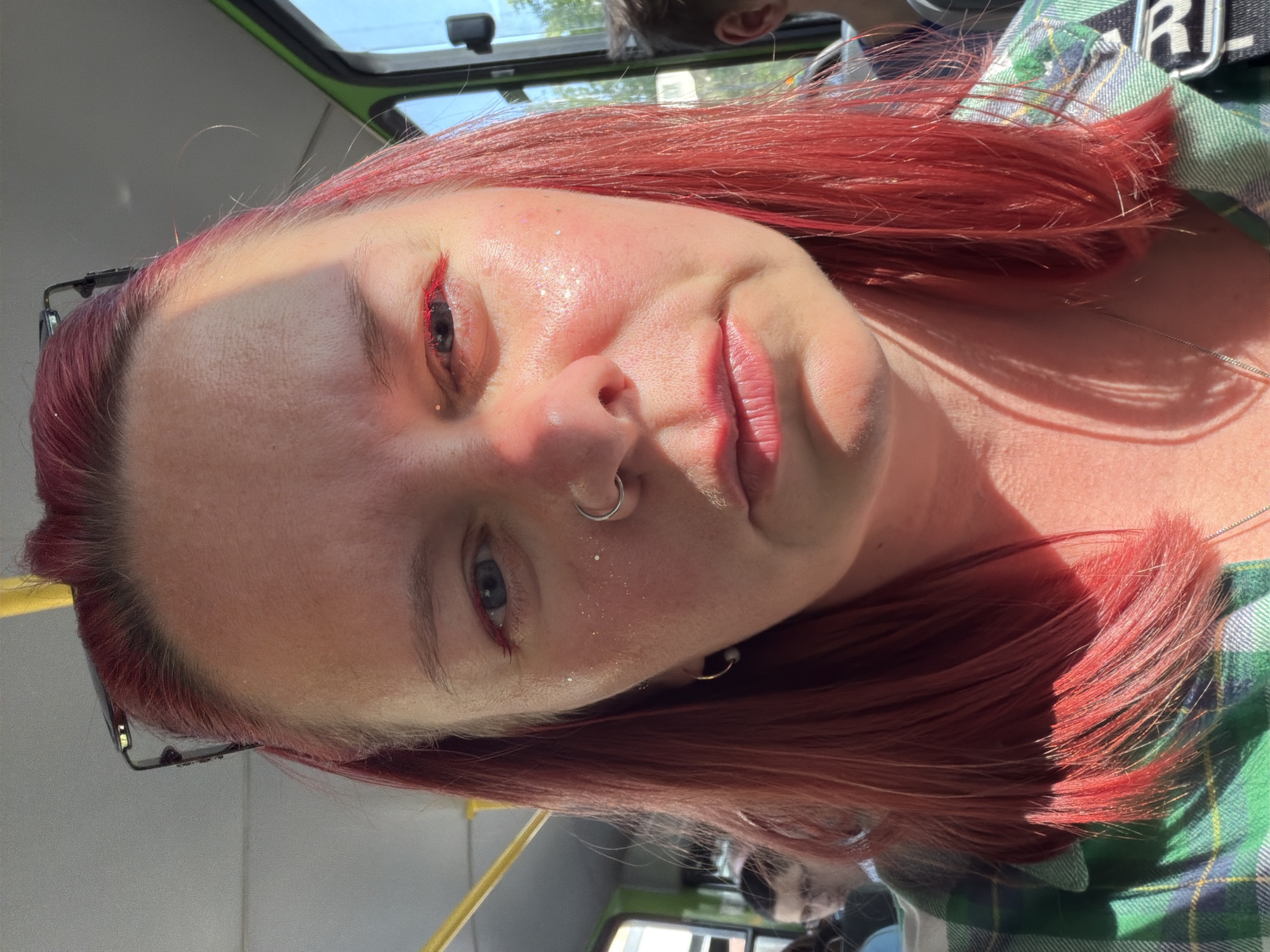 A close-up shot of a person with red hair on a bus, likely in Chaikovsky, Russia, given the request. The person has pale skin, red eyeliner under their eyes, a nose ring, and glitter on their face. They are wearing glasses pushed up on their head. The sun is shining brightly, creating harsh shadows on their face. They are wearing a green and blue plaid shirt. Other people are visible in the background, out of focus. A yellow pole is visible within the bus interior.
