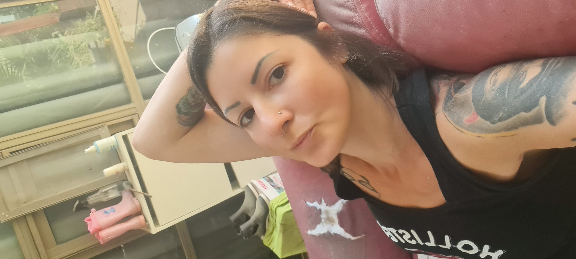 A woman is captured in a close-up, slightly tilted selfie, reclining indoors against a red, worn cushion, possibly a sofa armrest. She has dark hair, dark eyes, and a neutral expression, looking directly at the camera. Her left arm is bent, with her hand near her head, revealing a tattoo on her upper arm. Another large, dark, intricate tattoo is visible on her right arm, and a smaller one on her chest.
She wears a black tank top with partial white text visible, reading "HOLLISO...", likely "HOLLISTER". She has a small stud piercing on her left nostril and a ring piercing on her right, along with a silver-colored, star-like earring in her right ear.
The background features a window with light-colored frames, through which green foliage and reflections are visible, indicating daytime. Below the window, a light-colored shelf or cabinet holds several bottles and sprayers, a green cloth, and some pink items, possibly small boots or gloves. A book or magazine with the word "YOON" is also on the shelf. The red cushion she leans against shows a distinct white, damaged patch or design. The scene suggests a casual indoor setting within the Lachish Regional Council, Israel, with a view of outdoor greenery.