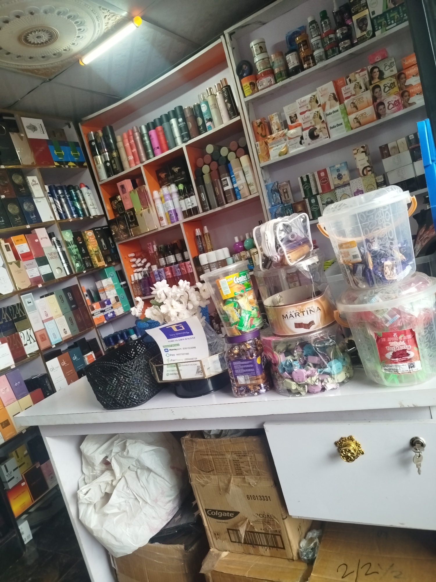 The image depicts a retail setting, likely a small shop or stall, with shelves stocked with various products. The items on display include perfumes, cosmetics, toiletries, and packaged snacks or sweets in plastic containers. There are also some boxes and bags, suggesting a storage or inventory area. The overall impression is of a well-stocked store. The ceiling features decorative molding and fluorescent lighting. In the lower part of the image, a white drawer with a gold handle and a silver key lock is visible, along with several cardboard boxes. The scene is brightly lit, and the merchandise is arranged in an organized manner on the shelves.
