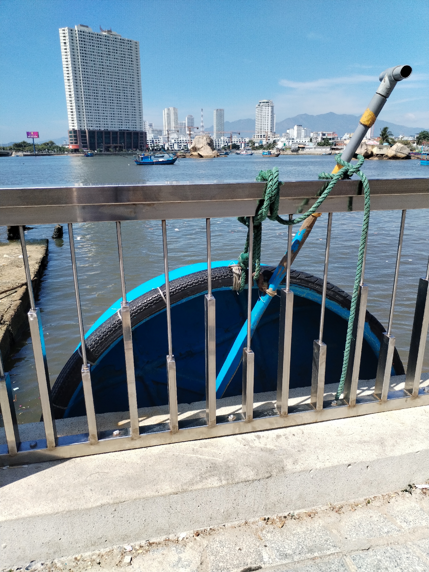A bright, sunny daytime scene shows a body of water in front of a city skyline. In the foreground, a stainless steel railing with vertical bars separates the viewer from the water. A blue boat with a black tire rim is tied to the railing with green ropes and a wooden pole extending upwards from the boat. The boat appears to be partially submerged. The water is choppy and a concrete walkway is visible in the lower part of the frame. In the background, a modern, white high-rise building dominates the skyline, with other smaller buildings and mountains visible further in the distance. The sky is clear and blue. The scene is likely located in Bac Nha Trang Ward, Vietnam, given the architectural style and the presence of a coastal city. There are no people visible in the frame.