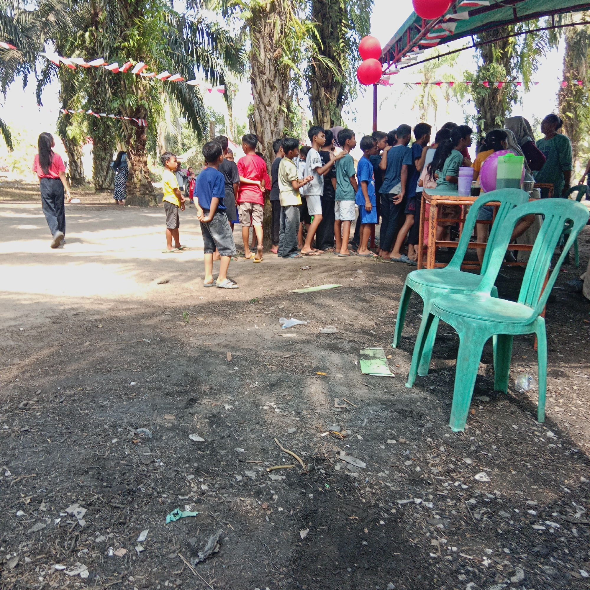 A group of people, mostly children and teenagers, are gathered outdoors under palm trees. Some are standing in a line, suggesting an event or queue. Red and white decorative flags are strung between the trees, and two red balloons are visible. In the foreground, two green plastic chairs are set up near a table. The ground is covered with dirt and debris, indicating an outdoor setting likely in a park or communal area. The overall scene suggests a festive or celebratory gathering. Based on the context of flags and balloons, it is likely this event is celebrating Indonesian Independence Day. The presence of palm trees and the general environment suggest a tropical location, possibly within the city of Dumai, Indonesia.