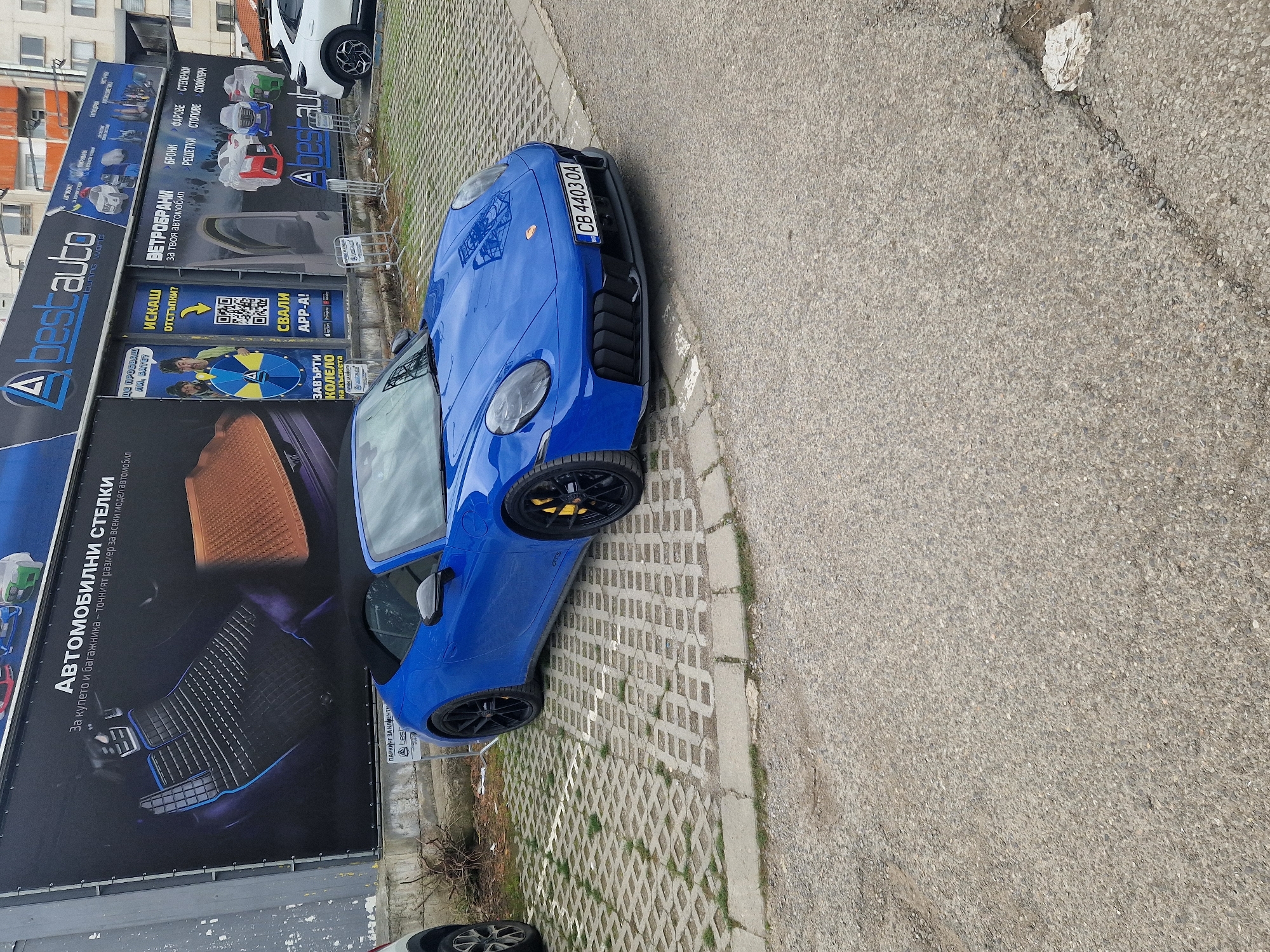 The scene is an outdoor shot of a blue Porsche GT3 parked on a gray interlocking brick surface, with a blurred gray asphalt road on the right. The car's license plate reads "CB 4403 OA". Adjacent to the car is a building with a large advertisement board featuring the text "Abest auto tuning world" and "АВТОМОБИЛНИ СТЕЛКИ" in Cyrillic, among other details of automotive products and services. Above the advertisement board, a white vehicle is partially visible. The environment includes an urban setting with a mix of building exteriors and street infrastructure.