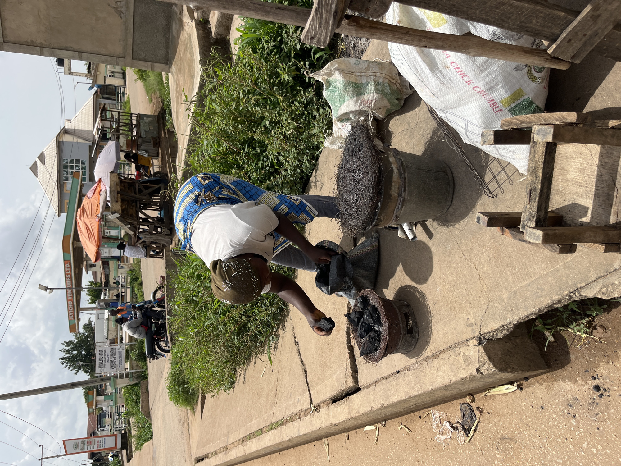 In Bukuru, Nigeria, a person wearing a blue and yellow patterned wrapper and a white shirt is engaged in an outdoor activity. They are holding a piece of charcoal, seemingly preparing to add it to a metal brazier filled with charcoal. The scene appears to be on a concrete path, with lush green vegetation growing nearby. In the background, there are buildings, some with pointed roofs, and a busy street with a gas station that has "JEVICHO VENTURES LTD." visible on its signage. Several motorcycles are parked along the side of the road, and people are seated at a roadside stall. A white sack with text that includes "A CHICK CRUMBLE" is also present, along with other bags and wooden structures. The sky is overcast, suggesting a cloudy day. The overall atmosphere suggests a community setting with commercial activity. Visible text includes "JEVICHO", "PRIVILEGE ABODE", "FOR CARE OF ORPHANS", "(PACOWF)", "& WIDOWS FOUNDATION", "SCHOOL FOR ORPHANS", "JEVICHO VENTURES LTD.", and "A CHICK CRUMBLE".
