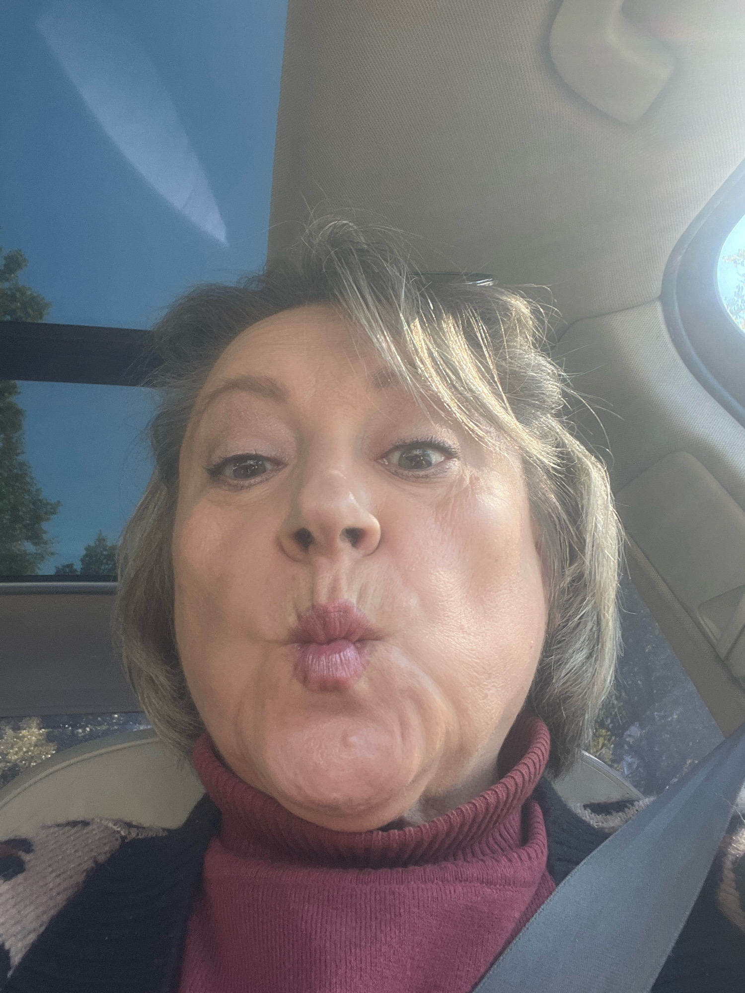 A woman is shown from the chest up, taking a selfie in a car. She has light brown hair and is wearing a red turtleneck sweater and a black cardigan with a leopard print collar. The car appears to be moving, as indicated by the sky visible through the sunroof and side window. The environment is a car interior, with the seatbelt visible across her shoulder. The time of day appears to be daytime, with sunlight filtering into the car. The location is not identifiable as Saint Petersburg, Russia.
