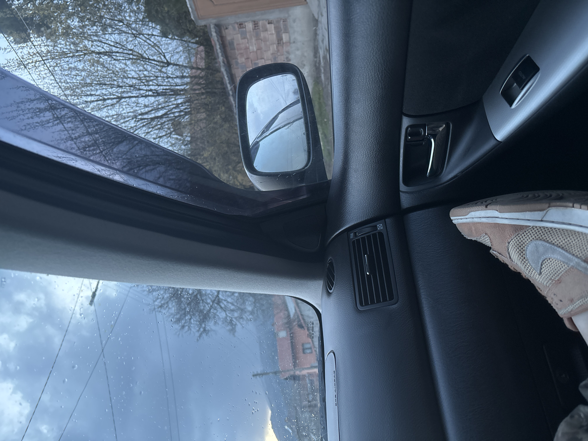 The image shows a view from inside a car, likely the driver's side. Raindrops are visible on the windshield. The side mirror is visible, reflecting a building and trees. The car's interior is mostly dark, with a silver door handle and window control panel. A portion of a light brown and white sneaker is visible near the center of the image. The car appears to be in Zlatitsa, Bulgaria, based on the visible building style and landscape in the side mirror and through the windshield. The specific street location cannot be determined from the image.