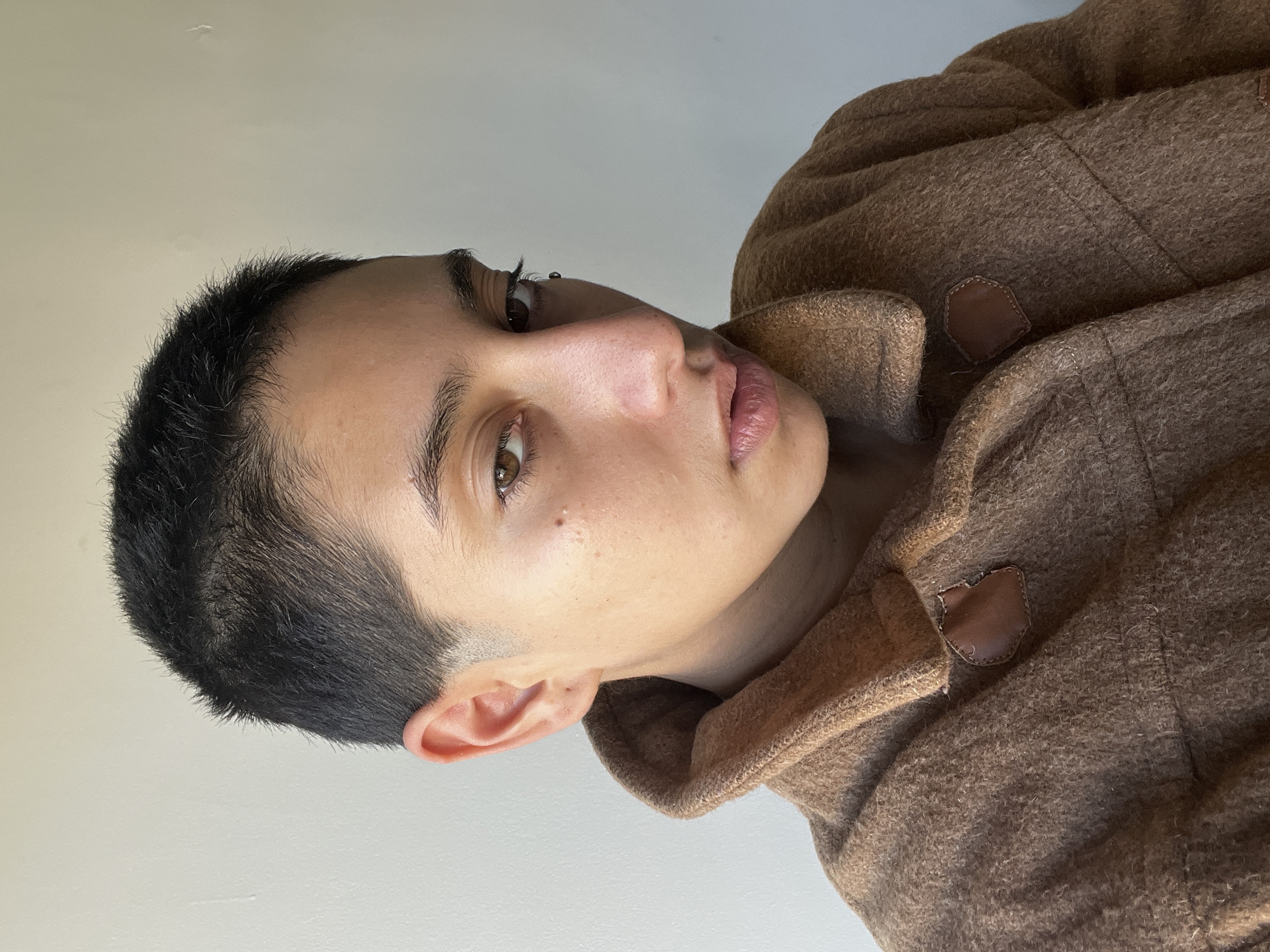 A medium shot depicts a person with short, dark hair and light skin, wearing a brown woolen coat. The person's face is turned towards the camera, with a slight upward angle. The background appears to be a plain, light-colored wall. The person has brown eyes and a visible piercing in their left ear. There is no visible information suggesting the location is Buenos Aires, Argentina.