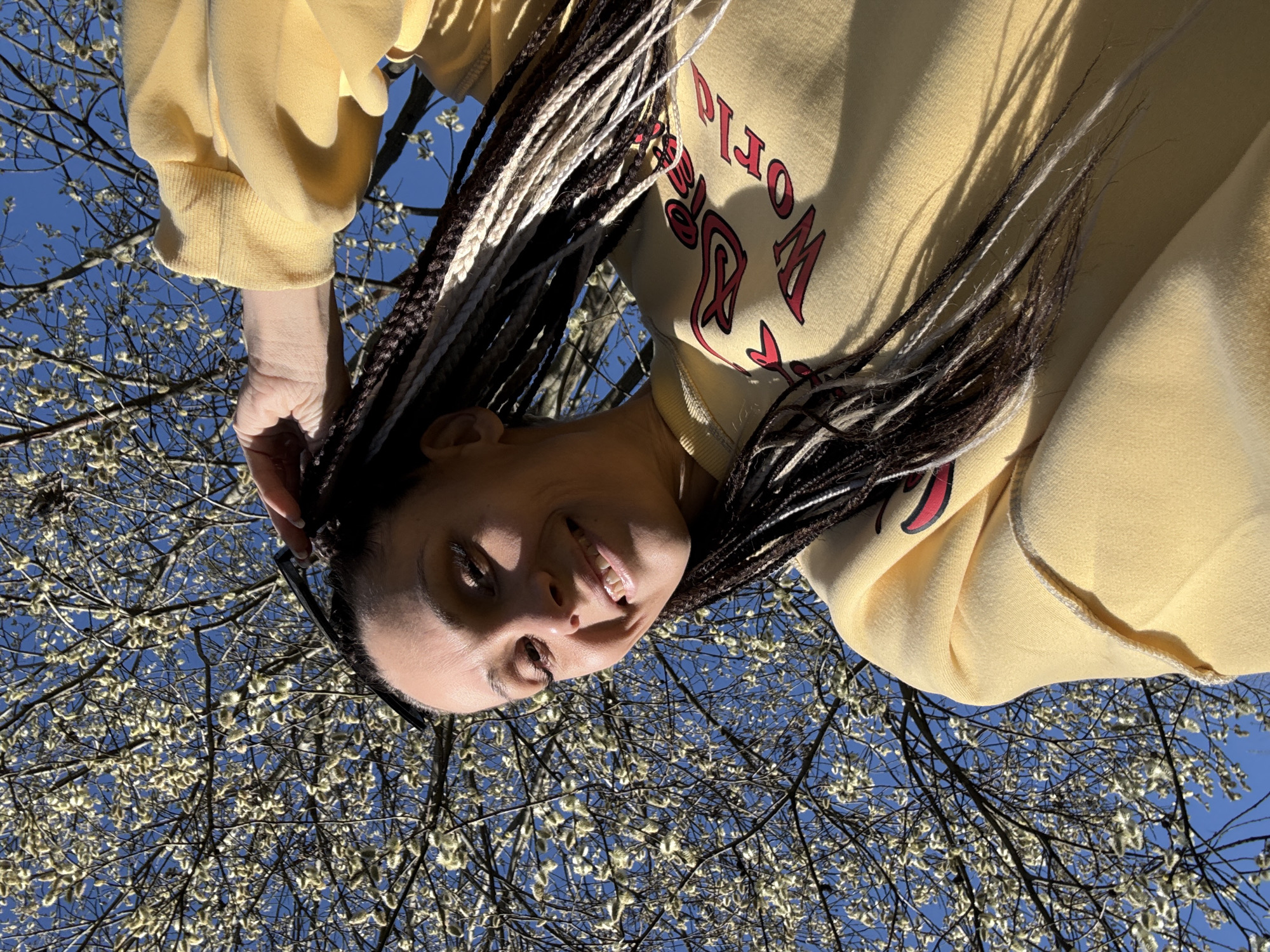 A woman with braided hair stands beneath a tree with emerging catkins against a bright blue sky. She wears a yellow sweatshirt with red lettering and holds her hand near her head, smiling slightly at the camera. The photograph is taken from a low angle, looking up. The location is not explicitly identifiable as Rusanovo, Russia, based on the visual information provided.