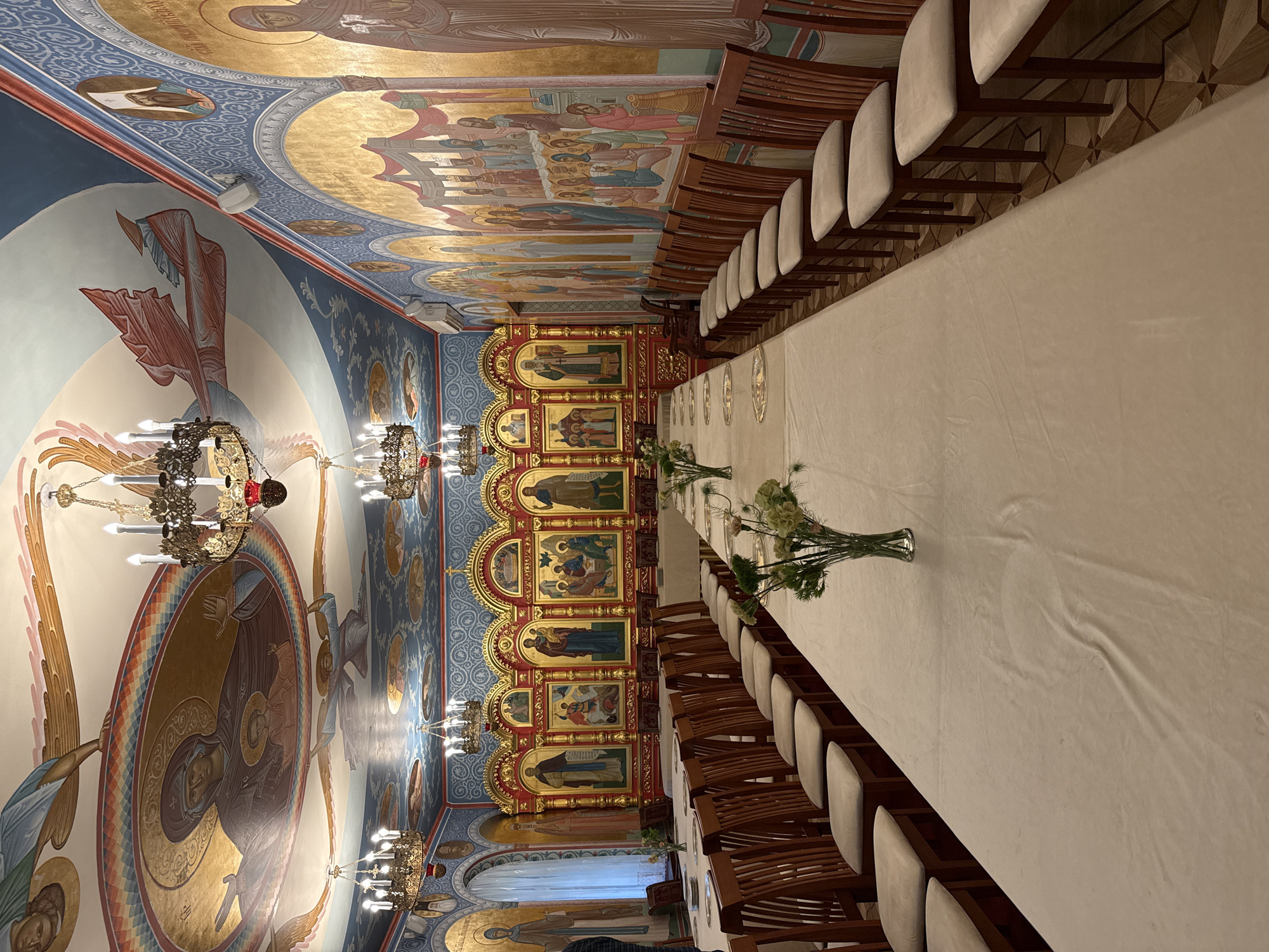 The image depicts the interior of a room, likely in a church or religious building. A long rectangular table covered with a white tablecloth stretches across the center of the frame, flanked by rows of chairs on either side. The chairs are upholstered with light-colored fabric and have wooden frames. Vases containing flowers are placed intermittently along the table.
The architecture suggests an Orthodox Christian context, with walls and ceilings adorned with religious iconography and murals. The walls feature panels of icons, detailed paintings of saints, and religious scenes. The ceiling is elaborately decorated with frescoes depicting biblical figures and narratives, with a color palette dominated by blues, golds, and reds. Chandeliers with multiple lights hang from the ceiling.
The overall scene gives the impression of a prepared space, possibly for a formal event or gathering within the religious setting. Given the architectural style and iconography, the location is plausibly within the city of Saint Petersburg, Russia, a city known for its historic Orthodox churches and cathedrals.