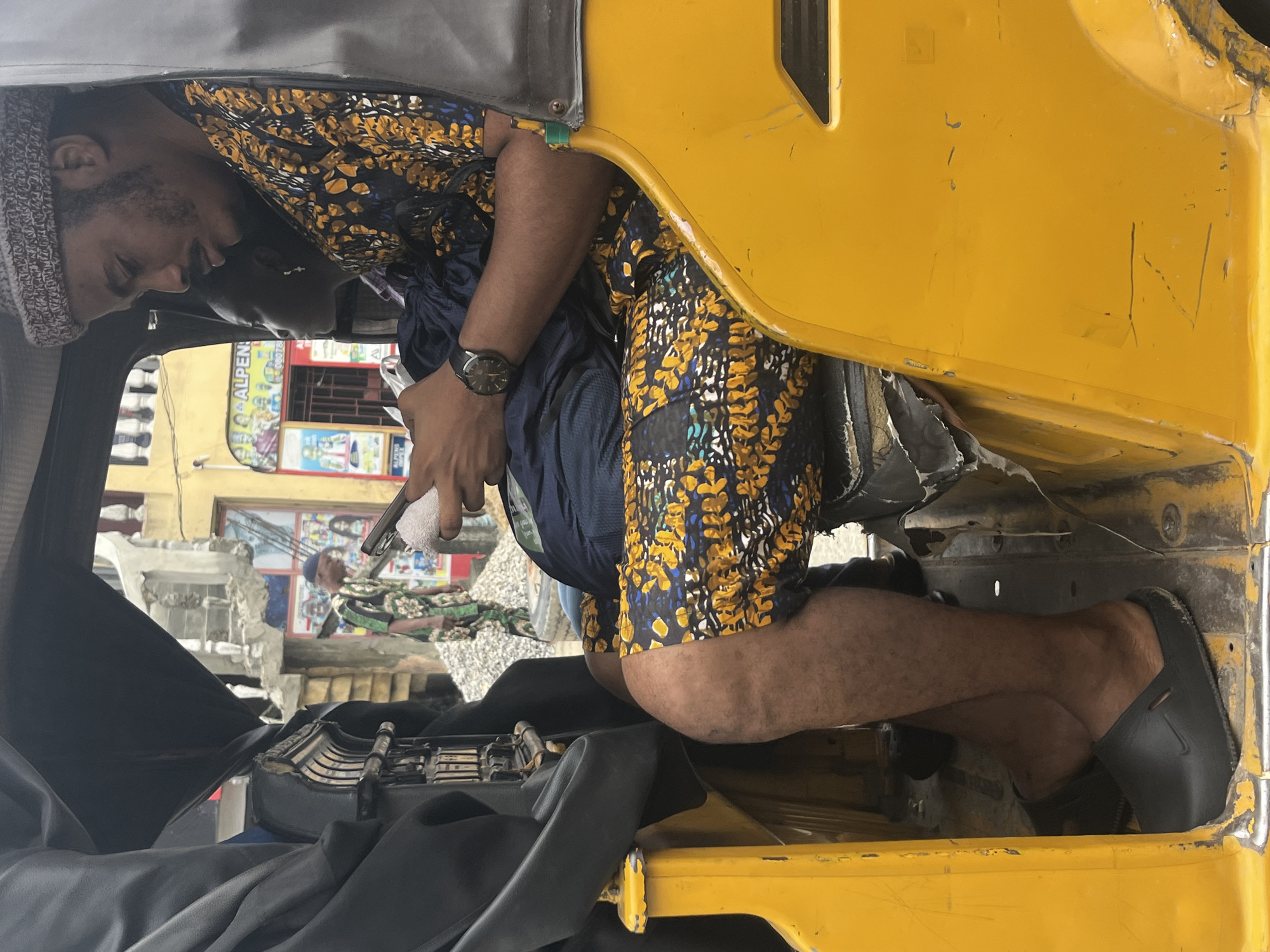 In Ado, Nigeria, a man wearing a patterned shirt and shorts sits in the passenger seat of a yellow auto-rickshaw. He wears a knit cap and a wristwatch, and holds a mobile phone. Beside him, a young girl is also seated. The background shows a street scene with buildings and other people. The auto-rickshaw is partially covered with a black tarp.