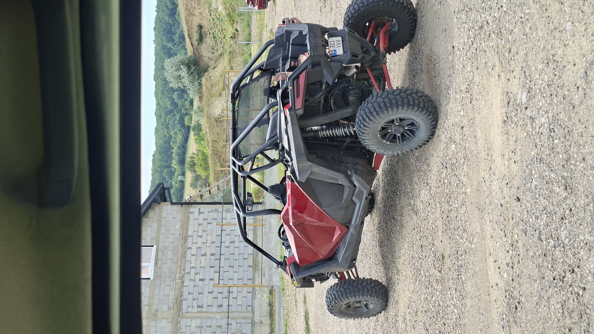 The image shows a red and black Polaris RZR off-road vehicle parked on a gravel surface. The vehicle has a roll cage, large tires, and suspension. In the background, there is a building under construction made of concrete blocks. Beyond the building, there are rolling hills covered in trees. A wooden ladder leans against the side of the unfinished building. The sky is visible in the upper portion of the image, and the scene appears to be outdoors in daylight. The vehicle has a license plate that reads "311 YXX".