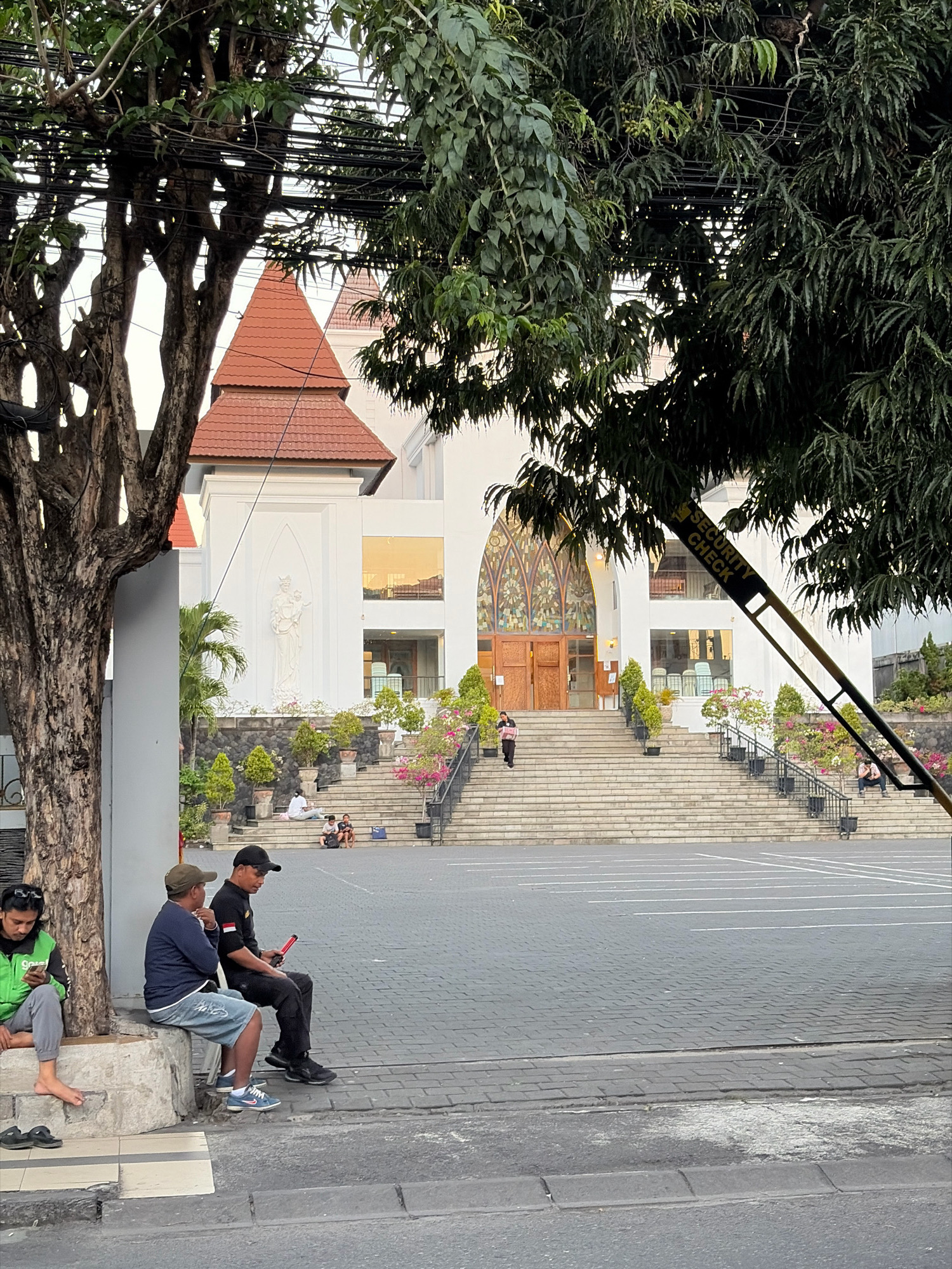 The image shows the entrance to a church in Kuta, Indonesia. In the foreground, a group of men are sitting on a low wall and on the pavement. One man is wearing a green jacket and looking at his phone, while another man in a blue shirt and denim shorts is talking to a man in a black shirt and black pants who is holding a red object. Behind them, a wide set of stairs leads up to the church entrance, which features a large stained-glass window and double doors. Several potted plants and trees are visible along the staircase and surrounding the building. A security check sign is visible in the upper right corner of the image. The overall scene appears to be a public area outside a religious building.