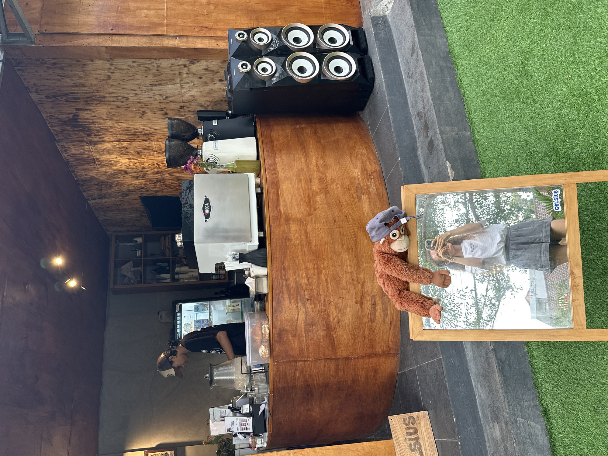 The image shows a street-level view of a cafe in Kuta Selatan, Indonesia. A barista is working behind a wooden counter, preparing drinks. Various coffee-making equipment, including a Wega espresso machine and grinders, are visible on the counter. In front of the counter, a plush orangutan toy with a hat is draped over a wooden easel holding a mirror. A young woman is taking a selfie in the mirror, with the cafe and some outdoor greenery reflected. To the right of the counter, a pair of tall, black speakers are positioned on the ground. The cafe has a rustic aesthetic with wooden walls and shelves. The overall scene suggests a casual and inviting atmosphere, possibly an outdoor seating area of a cafe in a tropical location.