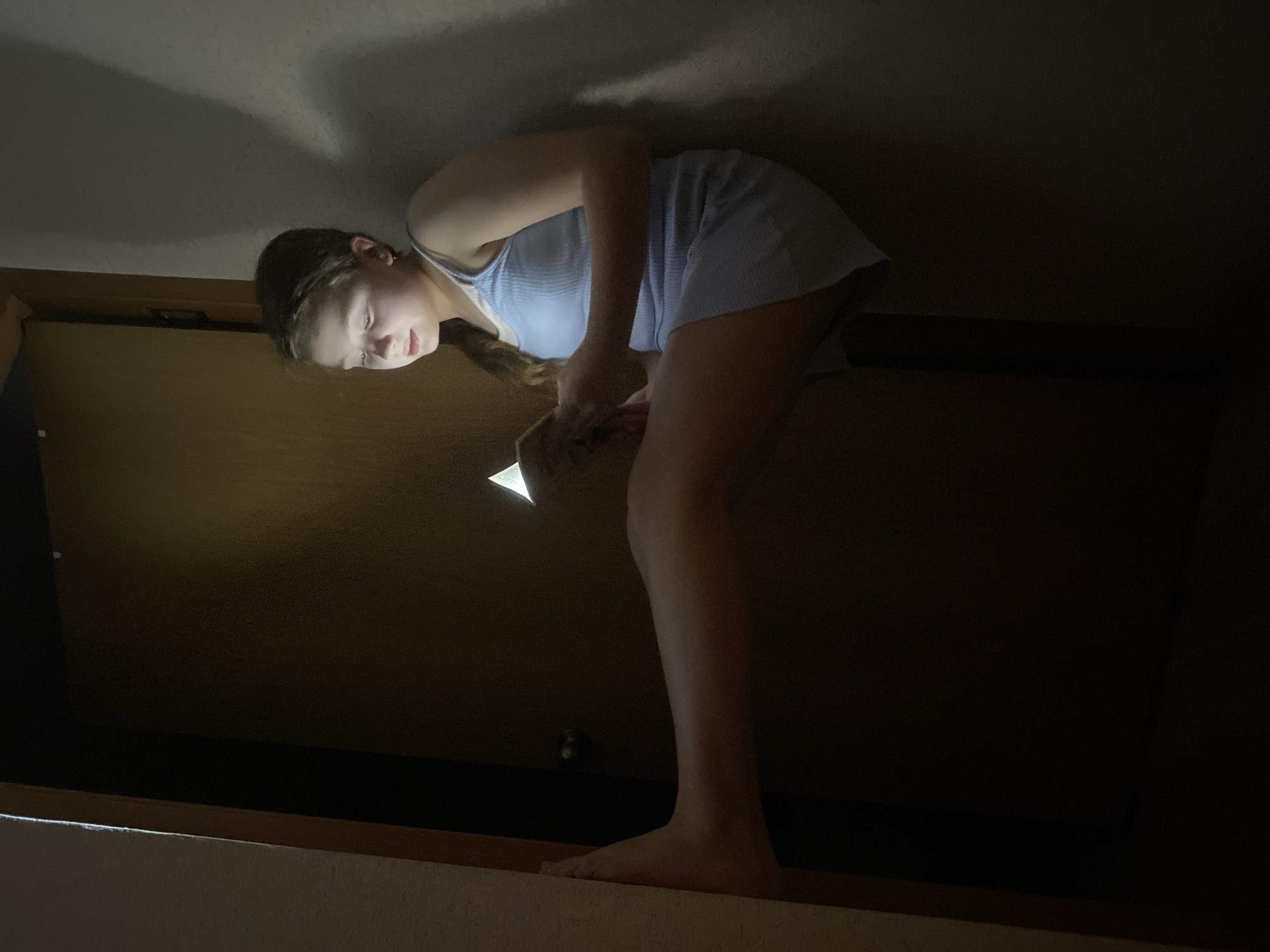 A young person stands in a dark, confined space, likely a closet or attic, holding a light source (possibly a phone) to illuminate an object. They are barefoot, wearing a light-colored, possibly blue or white, top and shorts. The environment is dimly lit, with only the immediate area around the person illuminated. The location is unknown.