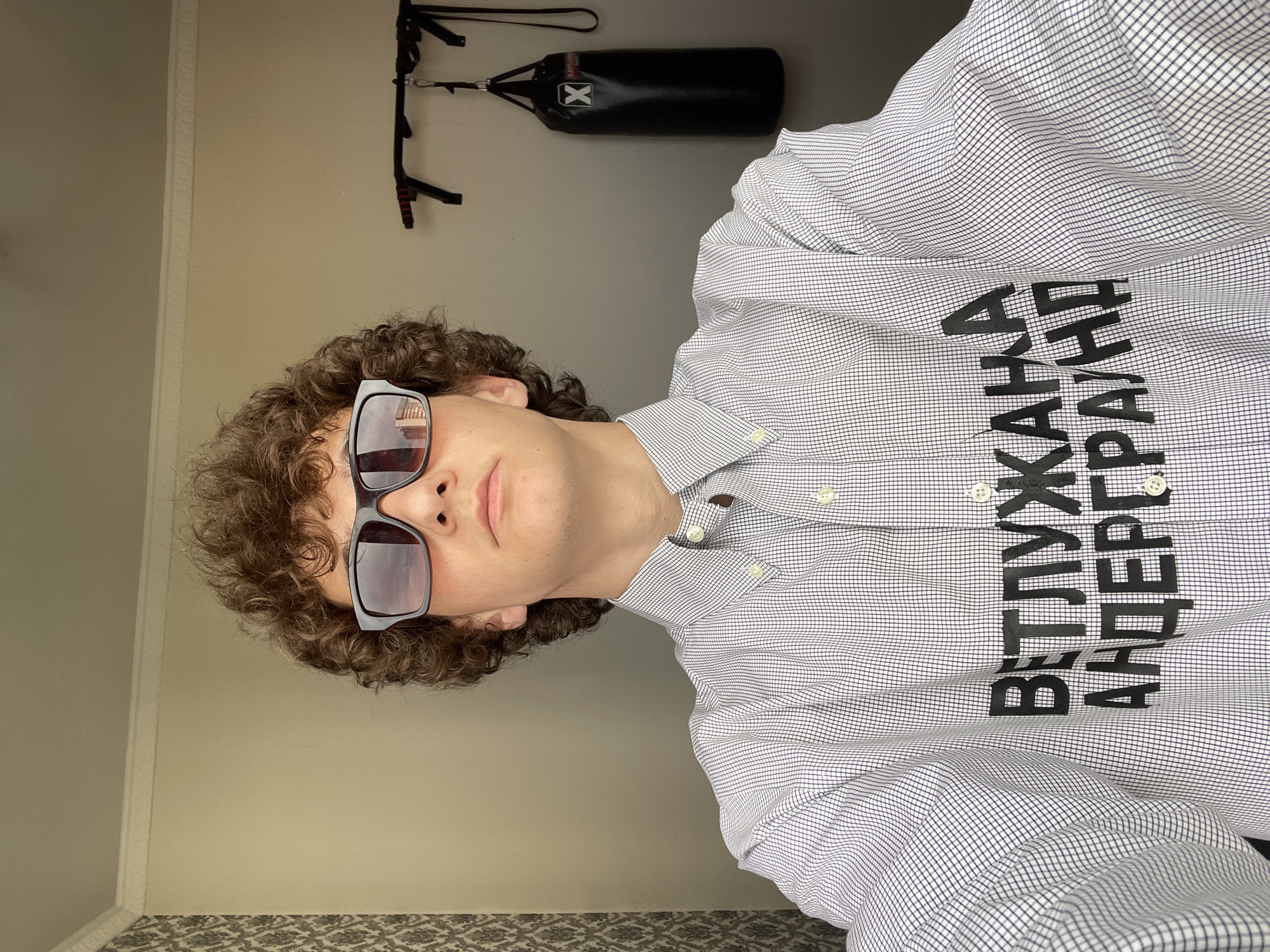 A vertically-oriented indoor shot showcases a person with curly brown hair, wearing sunglasses, and a checkered button-down shirt. The shirt features large black Cyrillic letters. Behind the person, the wall is a light beige color, and in the upper part of the frame, a black punching bag hangs from a black wall-mounted pull-up bar. The lower part of the frame includes a glimpse of a patterned floor or rug. There is no information to identify that the image was taken in the city of Krasnoyarsk, Russia.