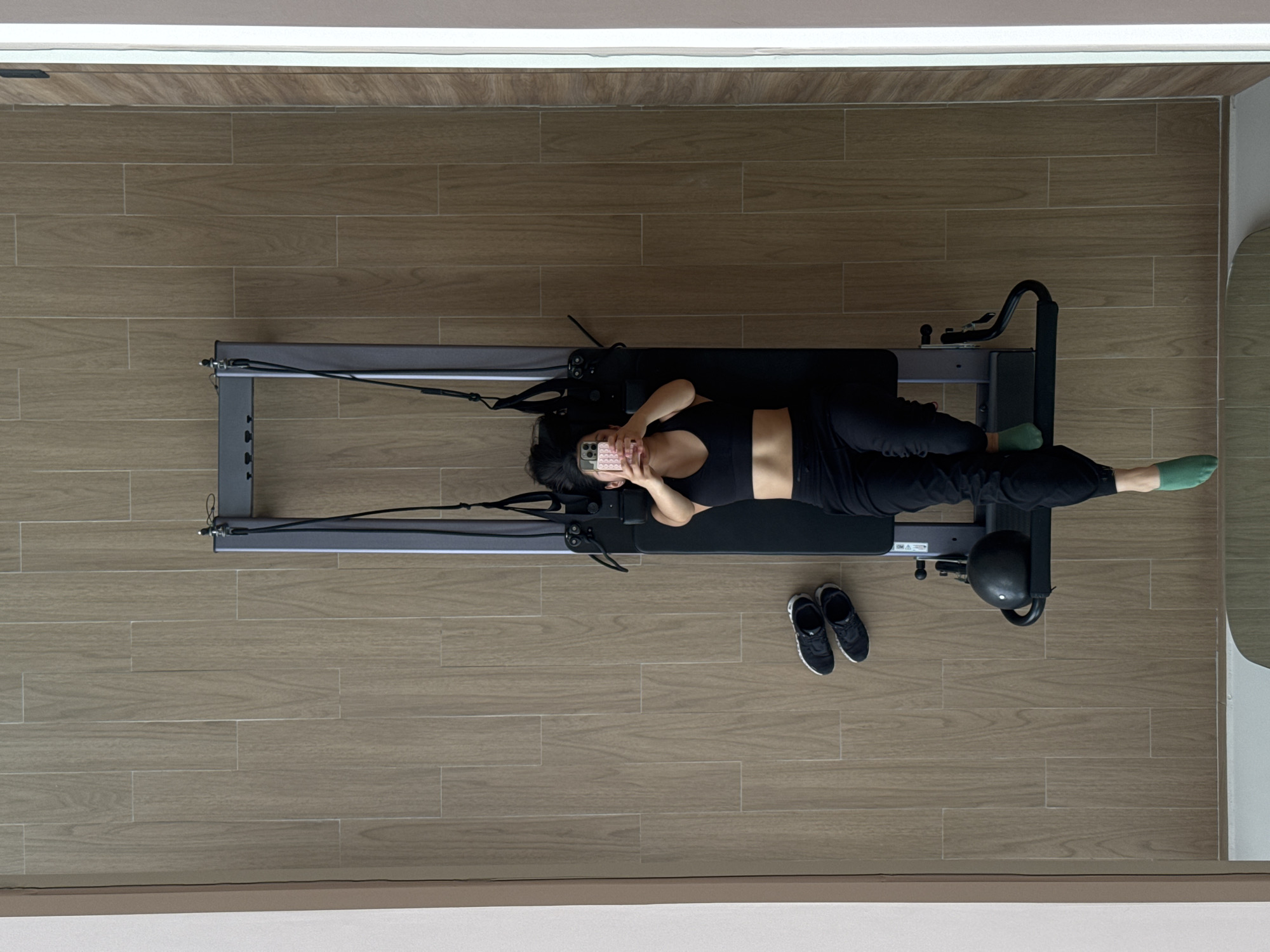 A woman in black athletic wear lies on a Pilates reformer machine in a room with light brown, wood-patterned flooring. Her feet wear green socks. She holds a phone with a pink case in her hands. A pair of black athletic shoes is next to the machine. The location is San Bernardino Tlaxcalancingo, Mexico.