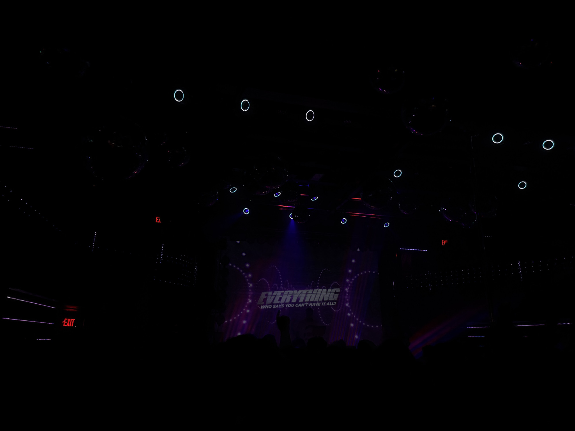 A darkened concert stage in a West Hollywood, United States of America venue shows a crowd facing a backdrop with "EVERYTHING WHO SAYS YOU CAN'T HAVE IT ALL?" printed on it. The stage is dimly lit with multiple circular stage lights and red exit signs, with some people visible in the audience.