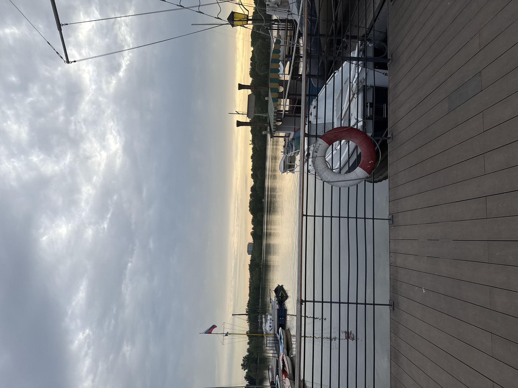 The image shows a waterfront scene in Moscow, Russia. The view is taken from a raised wooden deck, likely a dock or boardwalk, with a black metal railing. Beyond the railing, the water is calm, reflecting the sky. A row of boats is docked along the shore, some displaying the Russian flag. Further across the water, a treeline and a few buildings can be seen in the distance, under a cloudy sky. A life preserver ring is attached to the railing. The light suggests it is either early morning or late evening.