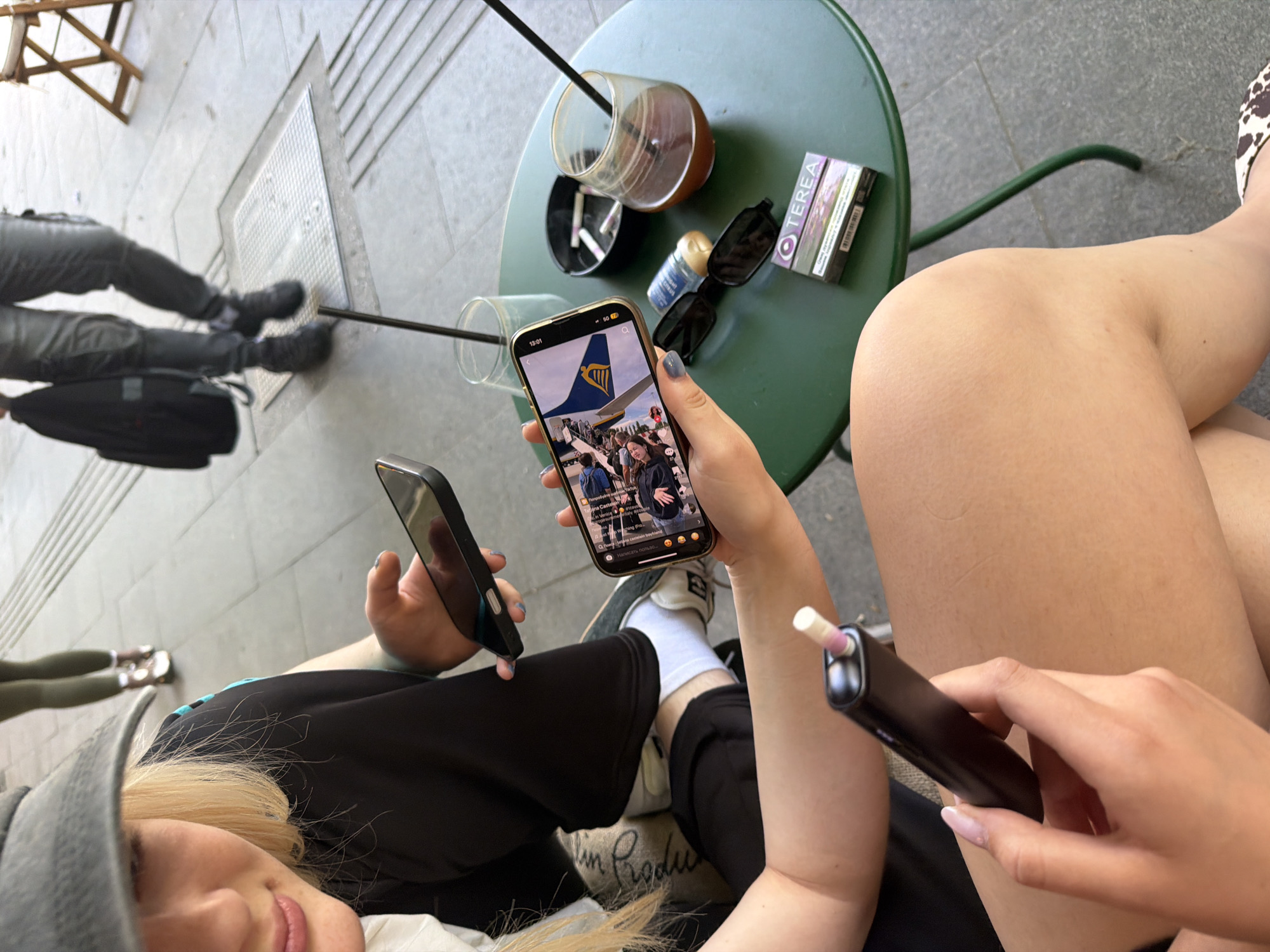 The image captures a close-up view of a person's hands holding a black electronic cigarette device and a smartphone, positioned in the foreground. The smartphone screen displays an image of people and a flag. In the background, a green circular table is visible, adorned with various items including sunglasses, a packet of Terea cigarettes, an ashtray with cigarette butts, and a glass of dark liquid with a straw. Another glass, partially obscured, is also present. The scene appears to be outdoors, likely a cafe or public seating area, suggested by the tiled ground and the presence of outdoor furniture. While the specific location of Belgrade, Serbia, is mentioned, no visual cues in the image definitively confirm this setting. The overall ambiance suggests a casual, relaxed moment.