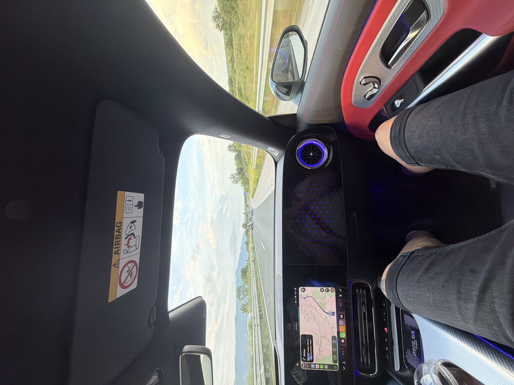 The image presents a first-person perspective from the front passenger seat of a modern Mercedes-Benz vehicle, traveling on a multi-lane highway in Bulgaria. The location is specified as the vicinity of Osikovitsa, a village in Lovech Province, supported by the navigation screen displaying destinations like Pleven and Lovech, and the highway designation A2.
The car's interior features a dual digital display system: the left screen shows a navigation map with a route highlighted, indicating "29 km" remaining to the current destination, a current time of 19:50, 350 km total distance remaining, and an ambient temperature of 21.5 degrees Celsius. The right screen displays a patterned array of illuminated blue Mercedes-Benz star logos against a dark background. A circular air vent on the right side of the dashboard is illuminated with a blue ring and a central starburst pattern. Below the main screens, a center console includes physical controls and a wireless charging pad or compartment with a custom mat bearing "PRESLAVA" and "МЕРЦЕДЕС" (Mercedes) in Cyrillic. A clear plastic cup with a lid and straw, containing a dark beverage, rests in a cup holder.
The passenger's legs are visible in the foreground, extended towards the dashboard, clad in dark shorts or rolled-up jeans, revealing knees and lower thighs. The passenger side door panel is accented with red upholstery featuring red stitching, silver-colored trim with a textured pattern, a silver door handle, and three silver seat adjustment controls. The passenger-side sun visor is raised, revealing a yellow "AIRBAG" warning label with universal symbols: a crossed-out rear-facing child seat, a depiction of an airbag deploying with a person seated incorrectly, and an icon indicating to read the owner's manual.
Outside the vehicle, a multi-lane asphalt highway is visible with other cars in the distance. The road is bordered by a green median, guard rails, and rolling hills covered with vegetation. The sky is partially cloudy with visible blue patches, indicating evening light consistent with the 19:50 time displayed on the navigation screen.