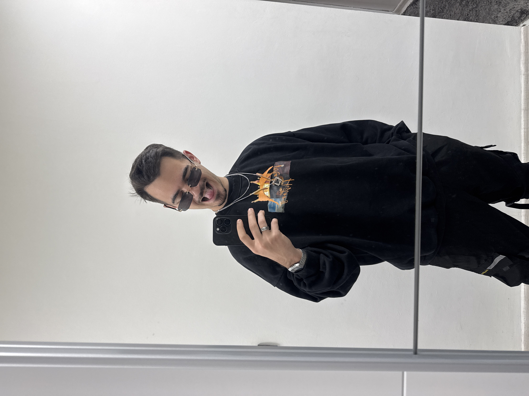 A man is taking a selfie in a mirror. He is wearing sunglasses, a black sweatshirt with a graphic on the front, black pants, a watch, and jewelry. The background consists of plain white walls and a section of light gray carpet in the upper right corner. The mirror is in a room with white walls, and there is no other specific indication of location within Sofia, Bulgaria.