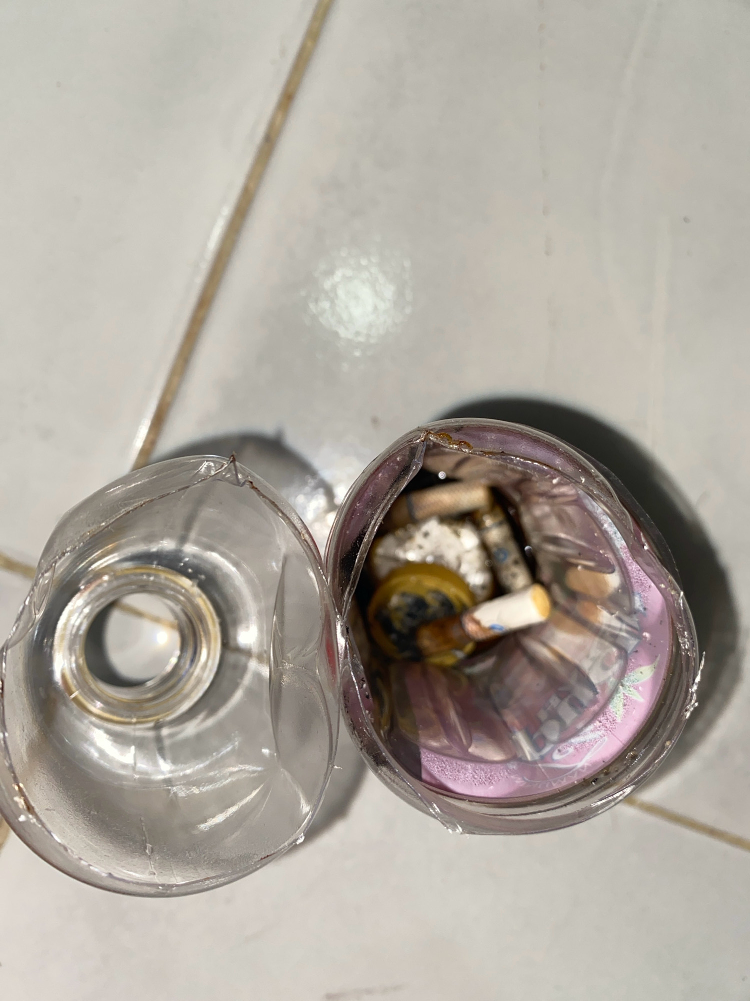 A cut plastic bottle lies on a tiled floor. The bottle has been cut into two pieces. One piece, the top, is empty and shows the opening. The other piece, the bottom, contains several used cigarette butts, a condom, and what appear to be folded banknotes. The scene is indoors, possibly in a domestic setting within Tanjung Pinang, Indonesia, given the tiled flooring and the presence of currency. The overall impression is one of neglect and disposal of waste.
