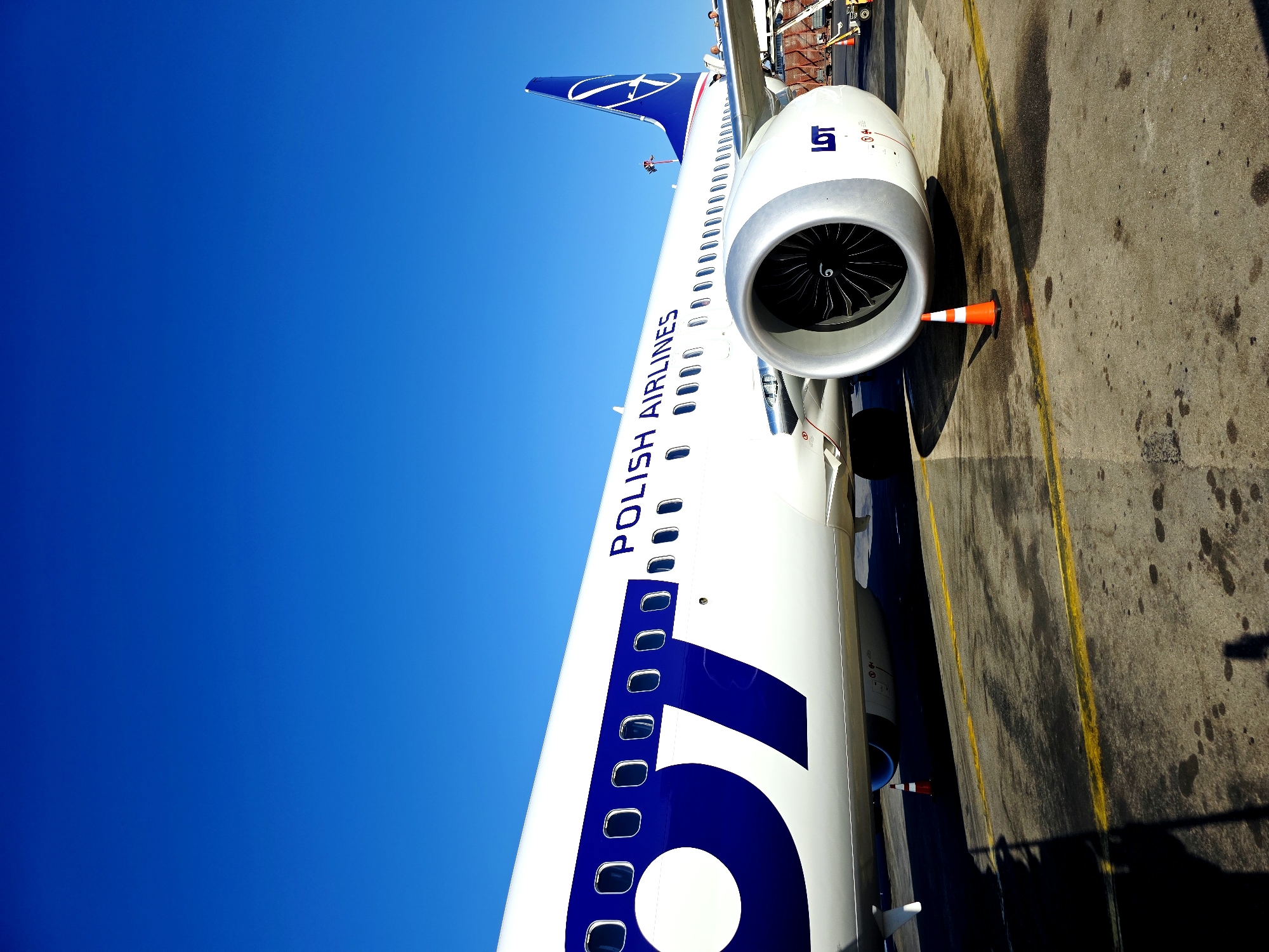 An airplane with the "POLISH AIRLINES" and "LOT" logos is parked on the tarmac under a clear blue sky. The aircraft is white with blue accents, including a large blue "OT" design on the fuselage and a blue tail fin with a white logo. A large jet engine is visible on the right side of the plane, with its fan blades clearly discernible. An orange and white traffic cone is placed in front of the engine. The tarmac is grey and marked with yellow lines. In the background, a baggage cart with red netting and a small yellow tug vehicle are visible. The image appears to have been taken from ground level, looking up at the airplane. The scene is consistent with an airport environment.