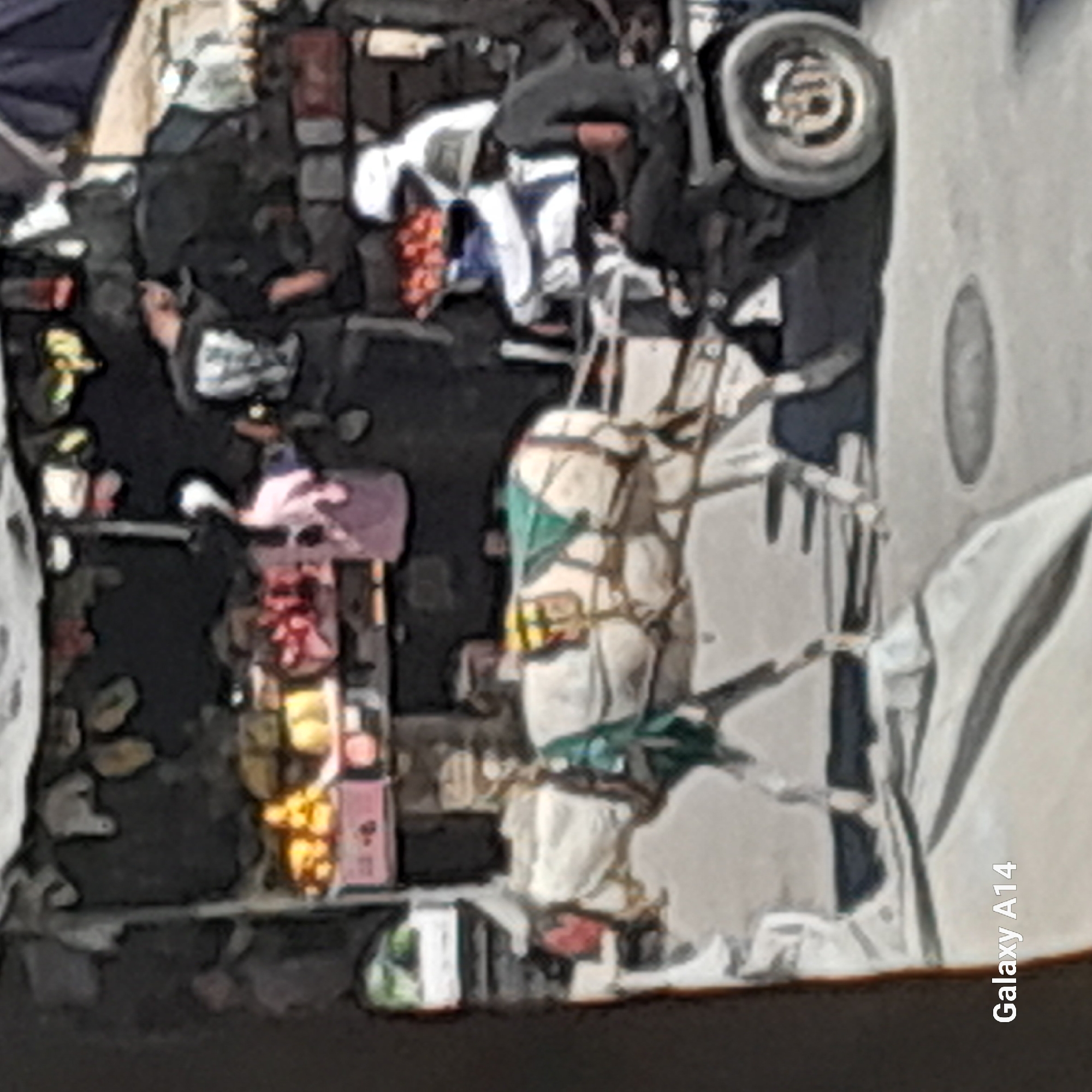 An aerial view captures a scene in Khan Yunis, Palestinian Territories, depicting a market or street setting with a horse-drawn cart. The cart is laden with various items, including what appear to be textiles and possibly food items, wrapped in white and green coverings. The horse pulling the cart is partially obscured. In the background, there are glimpses of people in dark clothing, some bent over or engaged in activities around the cart, suggesting a commercial or transport operation. The ground is a mix of dark, possibly dirt or paved surfaces, with some debris. To the right of the frame, a grey concrete structure is visible, possibly a wall or building facade, with a circular indentation. The overall image has a slightly stylized, almost painted quality due to image processing. A watermark, "Galaxy A14", is present on the lower right side.
