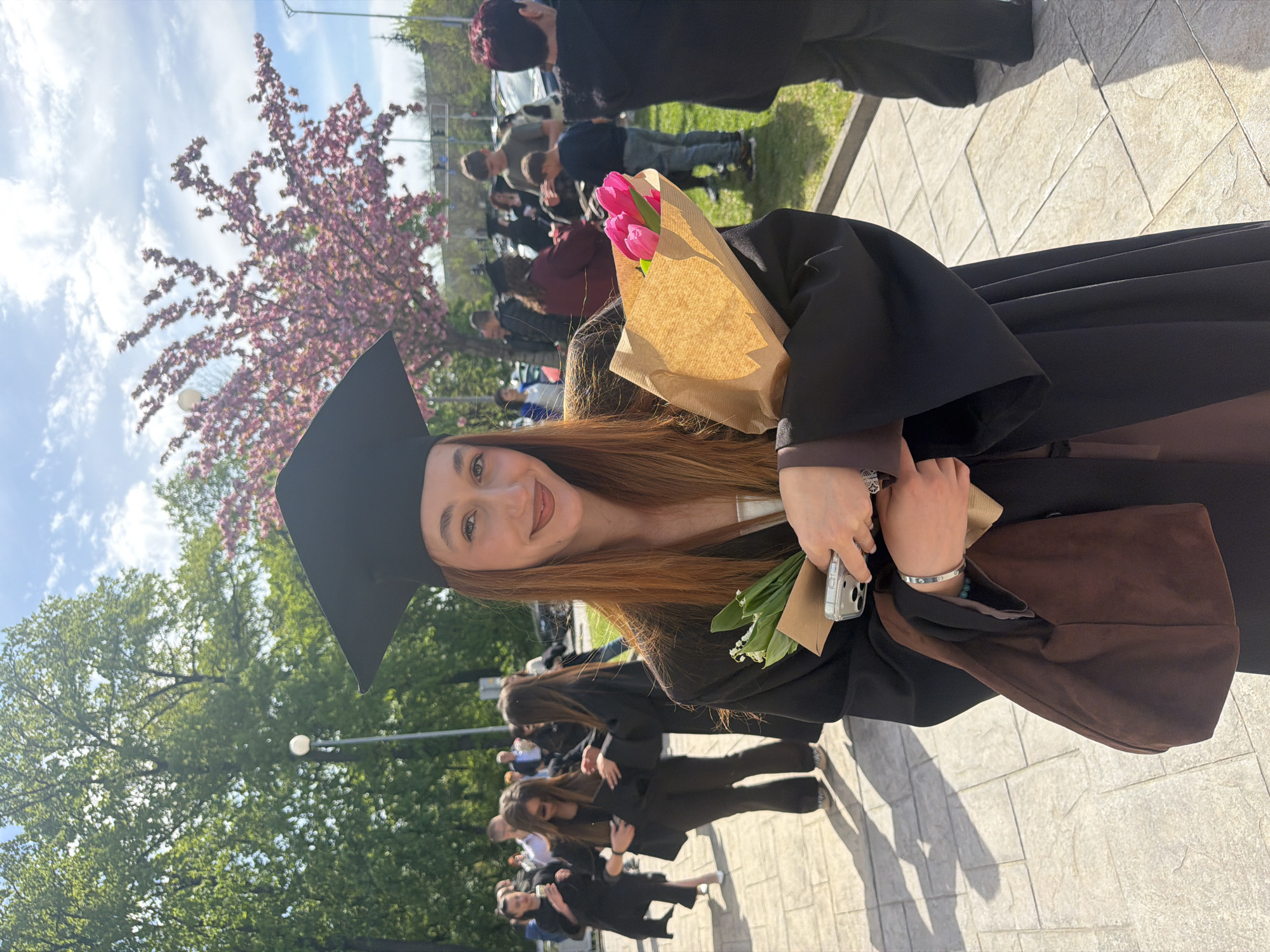 A young woman in a black graduation gown and cap is holding a bouquet of pink tulips. She is outdoors, likely in a park or campus area, with trees and other people visible in the background. The presence of a blossoming tree indicates it may be spring. Given the prompt, the location is assumed to be in Plovdiv, Bulgaria. Other graduates are visible in the background, suggesting a graduation ceremony or gathering.