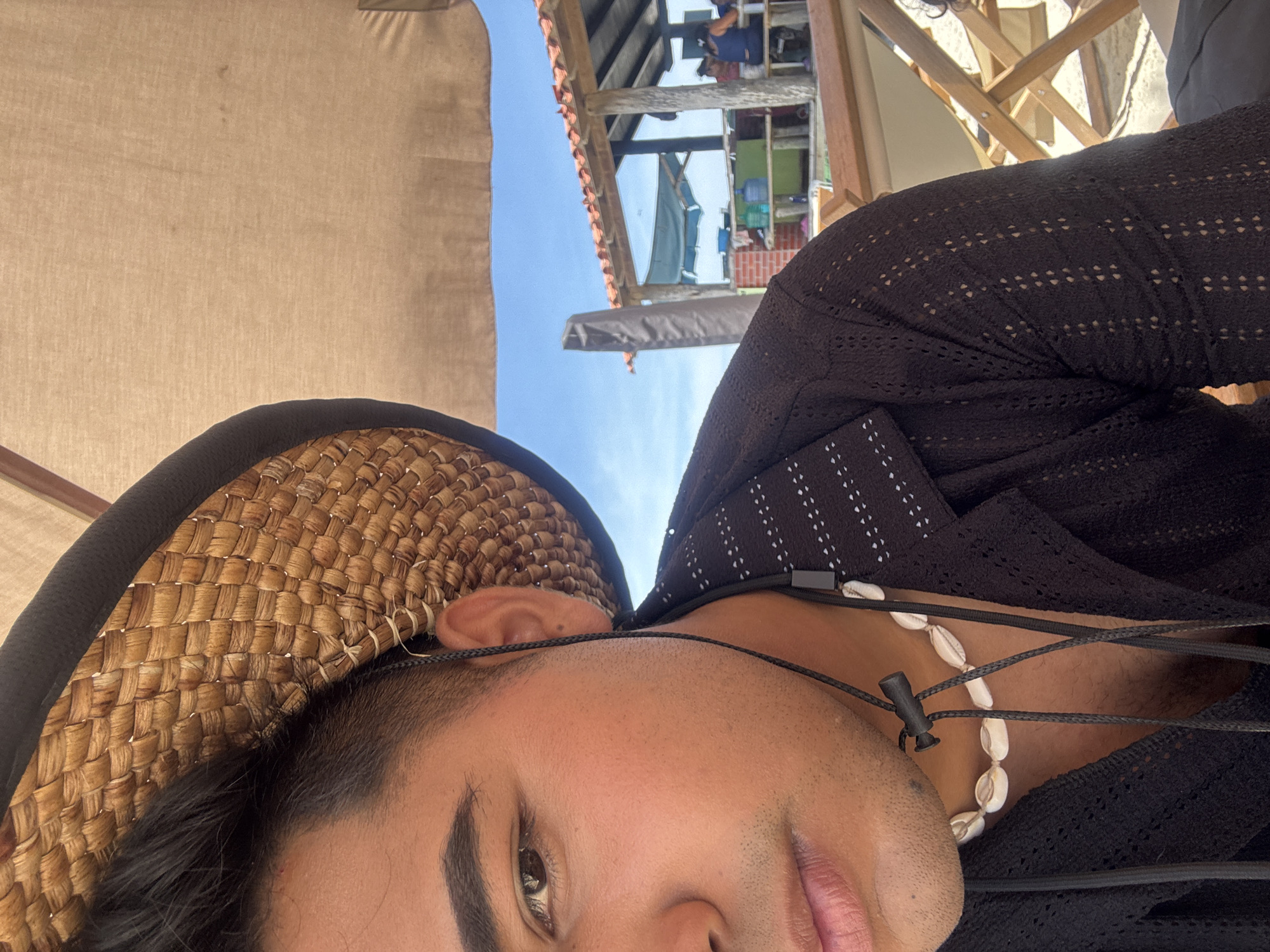 A low-angle, close-up shot features a person wearing a straw hat with a black brim and a black, open-weave shirt. They have a light-skinned complexion and are also wearing a necklace with white seashell beads. Parts of a wooden structure and a building with a red tile roof can be seen in the background, along with a glimpse of a blue sky. The location is suggested to be Porlamar, Venezuela.